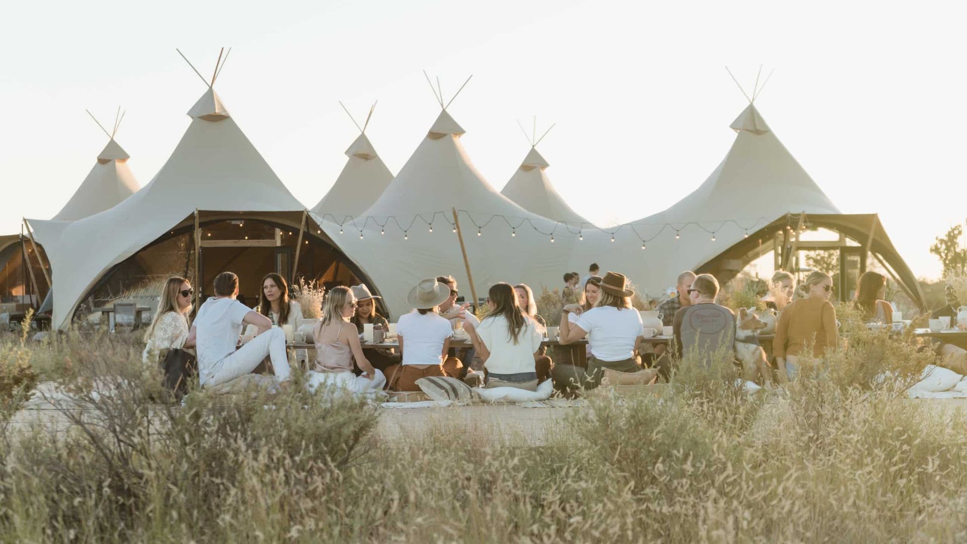 People gather around a low table in front of several large teepee-style tents in an outdoor setting.