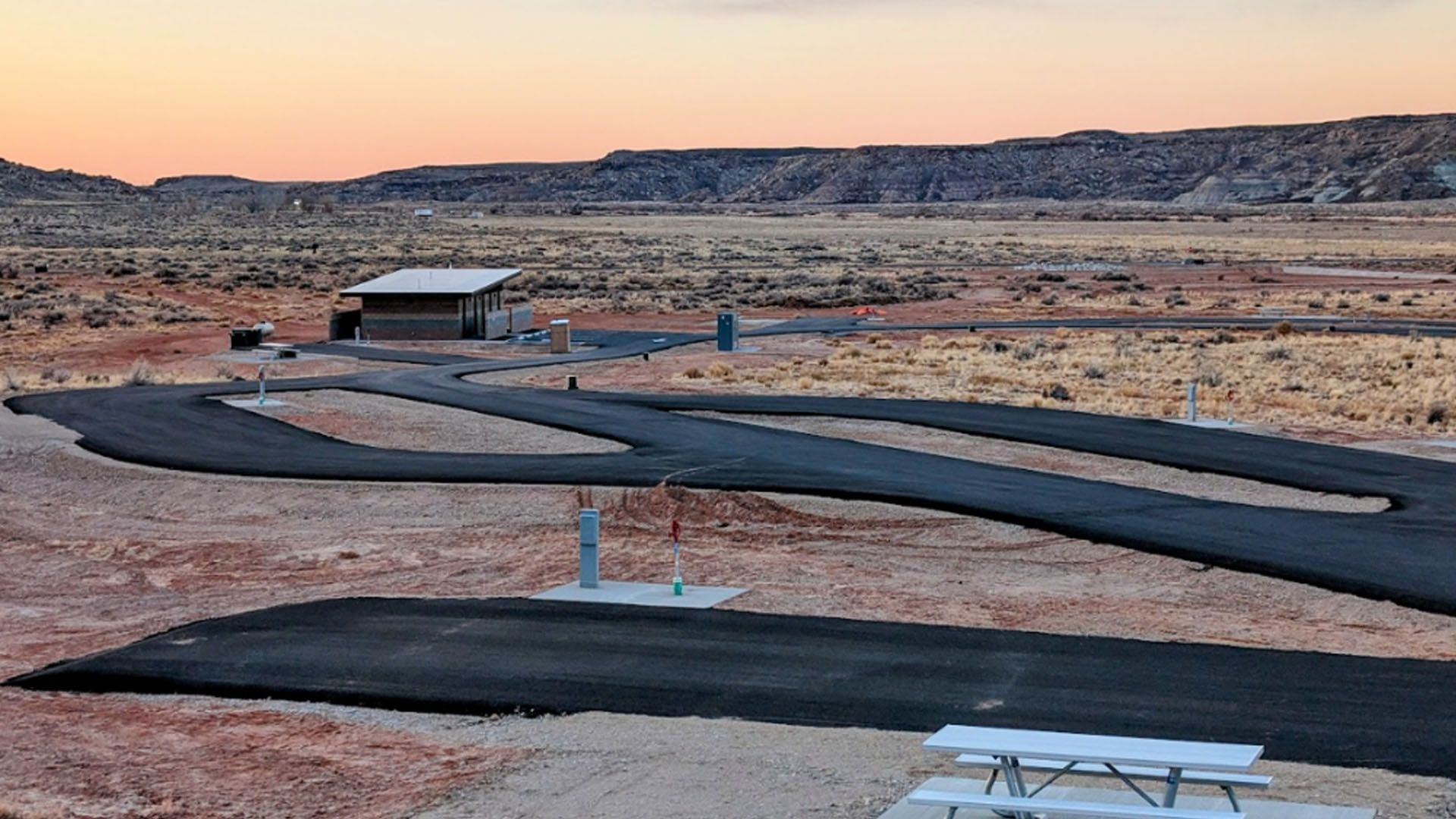 Desert campsite with paved roads, picnic table, and shelter under a colorful sunset.