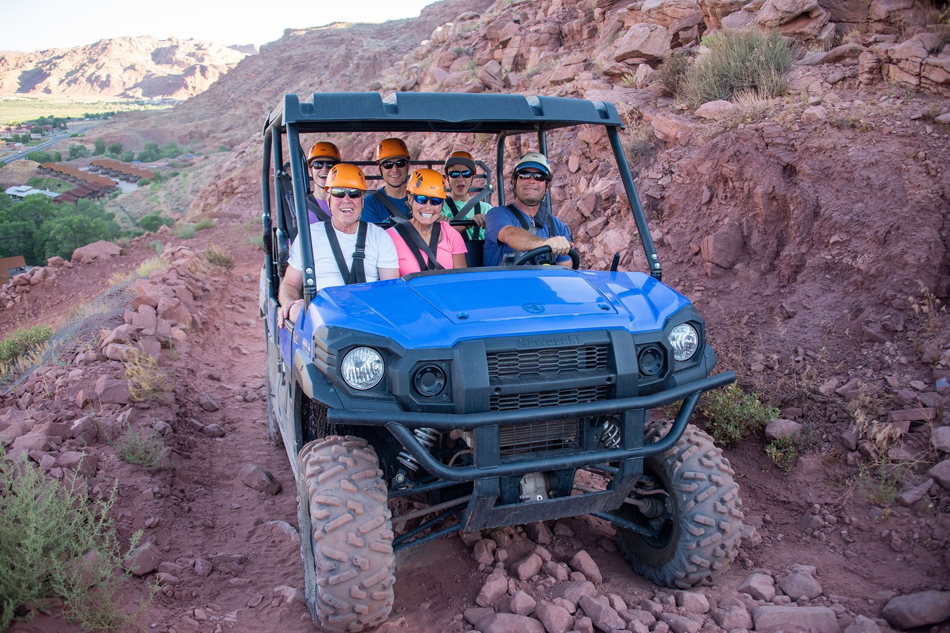 Blue off-road vehicle with five people wearing helmets on a red dirt trail in a canyon.