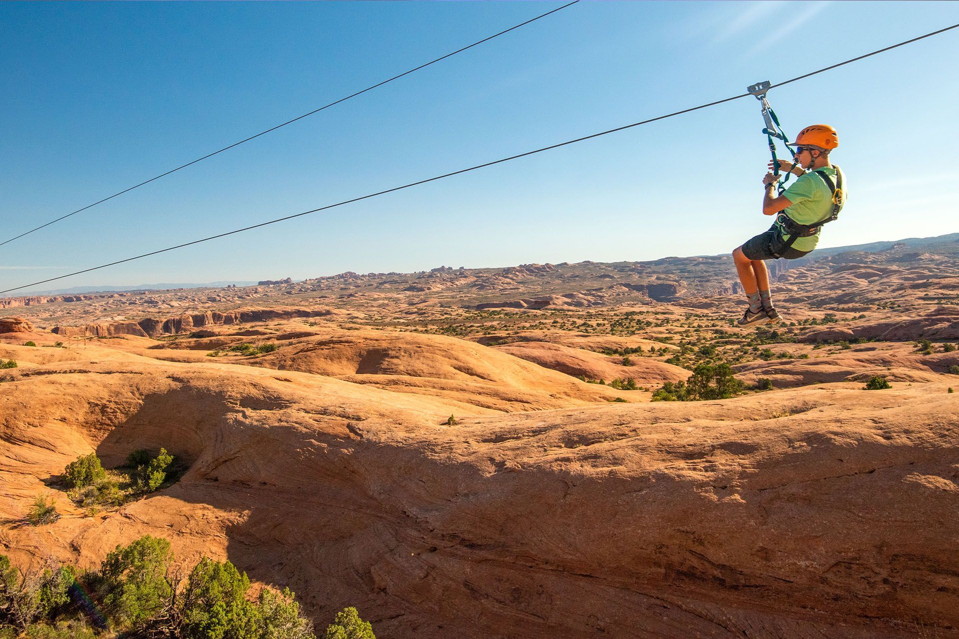 Person ziplining over a desert landscape, wearing a helmet and harness, against a blue sky.