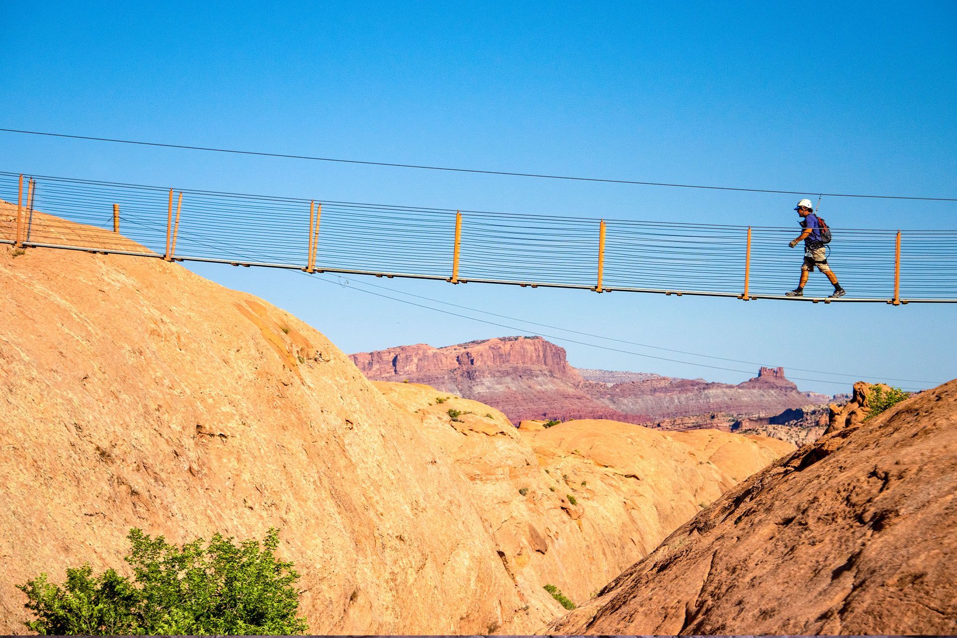 Person walks a suspension bridge over a desert canyon; red rock formations and blue sky.