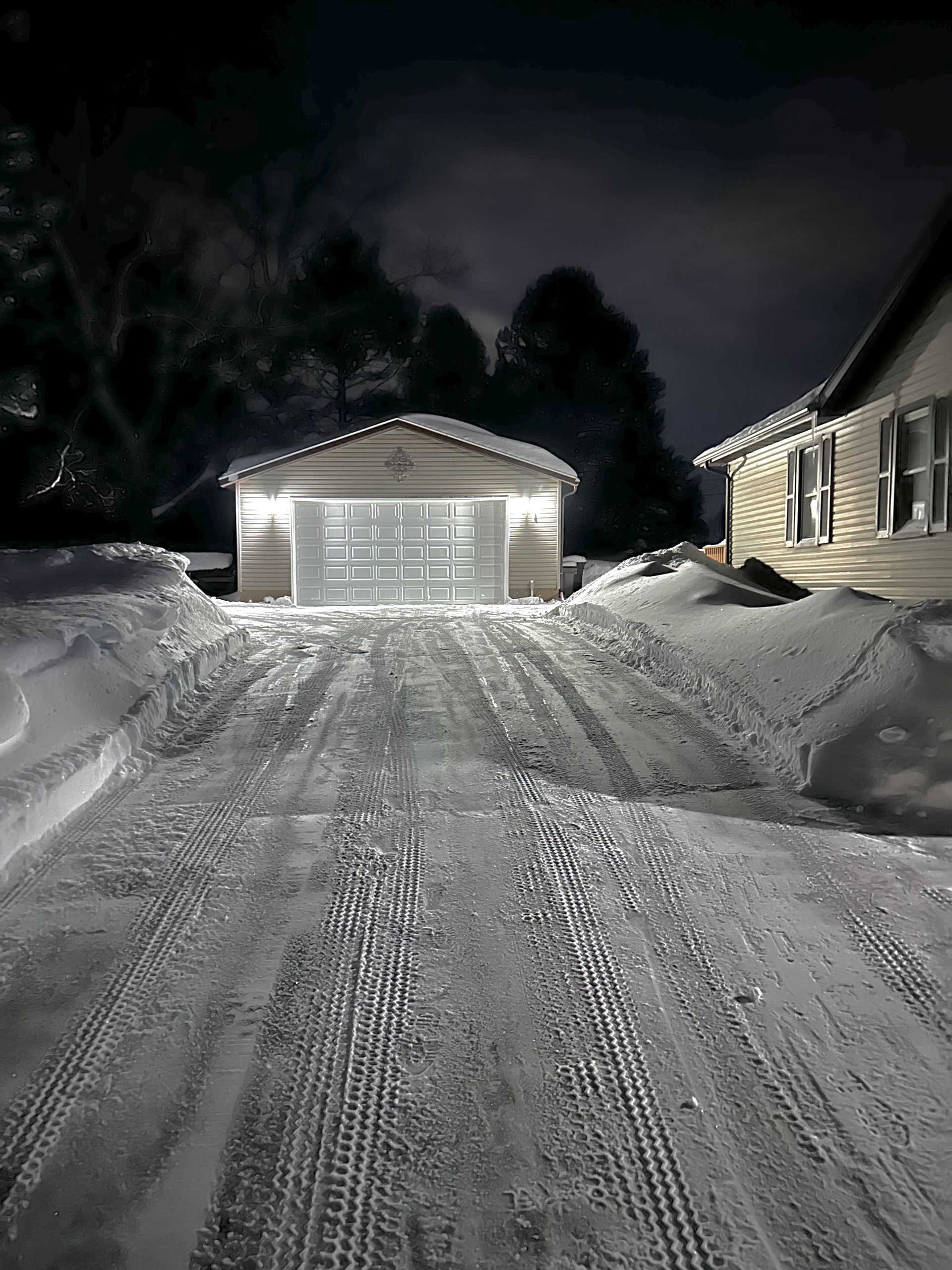 A snowy driveway leading to a garage and a house at night.