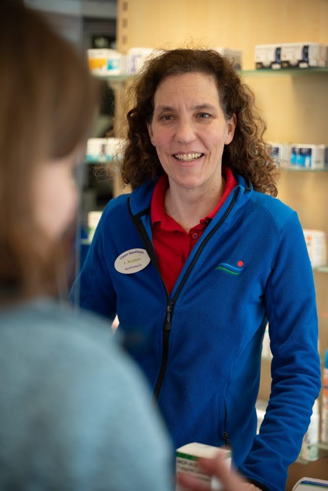 Eine Frau in einer blauen Jacke spricht mit einem Kunden in einer Apotheke.