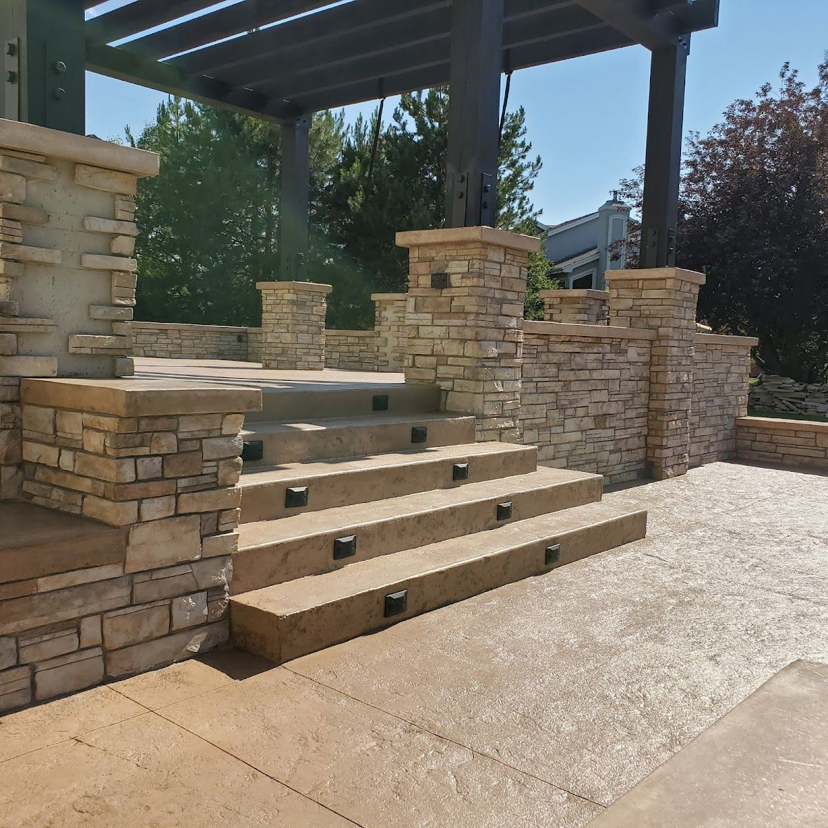 Stone steps lead up to a raised patio with a pergola, built with stone and wood, outdoors in sunlight.
