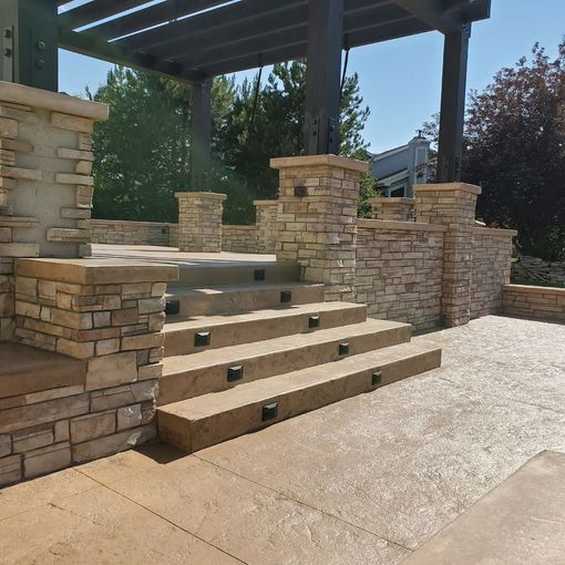 Stone steps lead to a raised patio under a pergola; set on a tan stamped concrete surface, with blue sky.