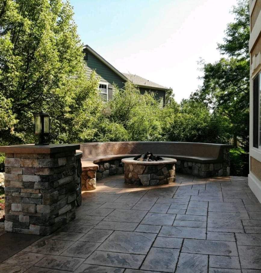 Stone patio with fire pit and built-in seating, near a house and trees.