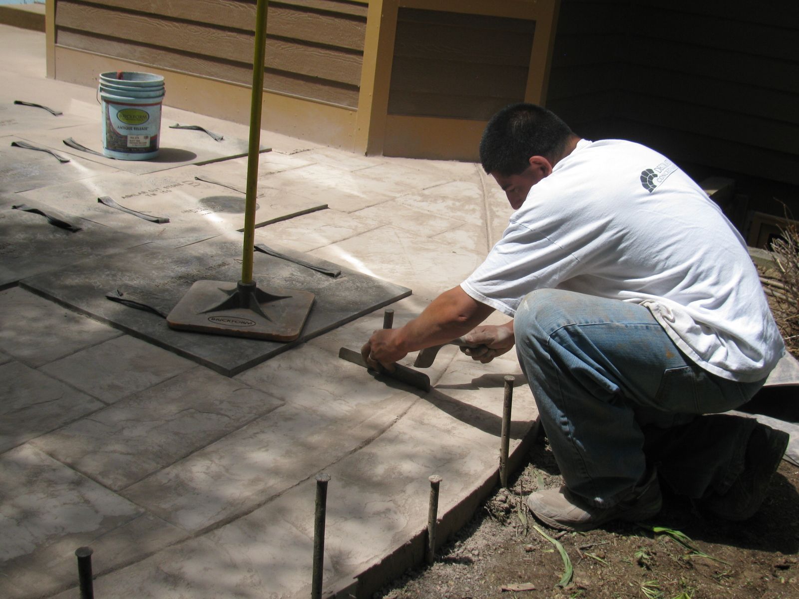 Man using tools to work on a concrete patio, near a bucket and a wooden structure outdoors.