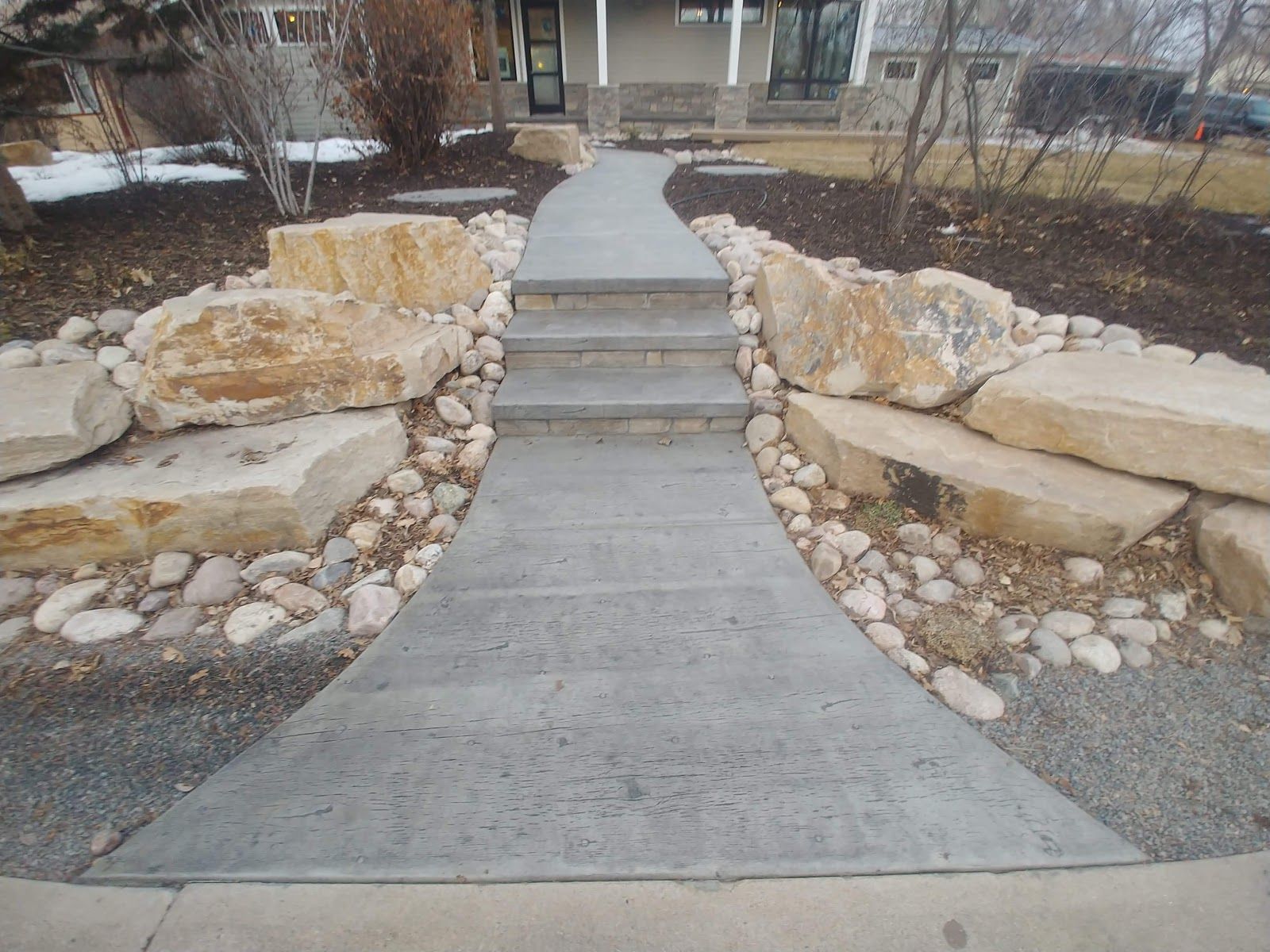 Concrete walkway with steps, bordered by large rocks and smaller stones, leading to a house.