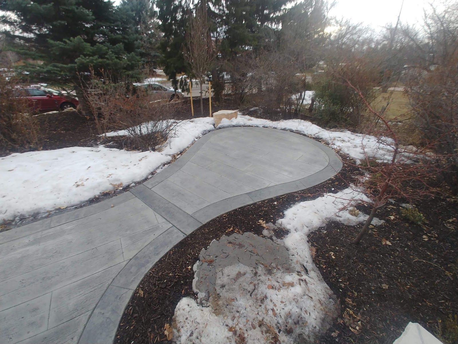 Curved concrete driveway and patio, edged with dark border, in a snowy landscape.