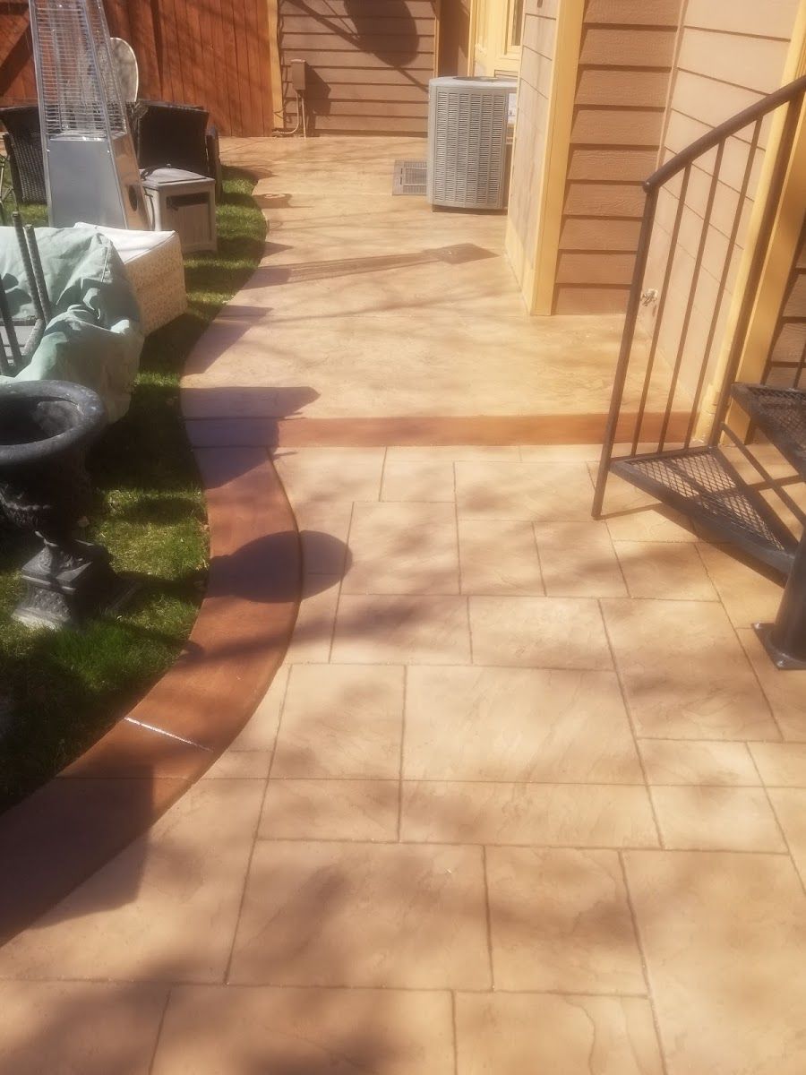 Tan patio with darker border next to grass and a building with metal stairs.