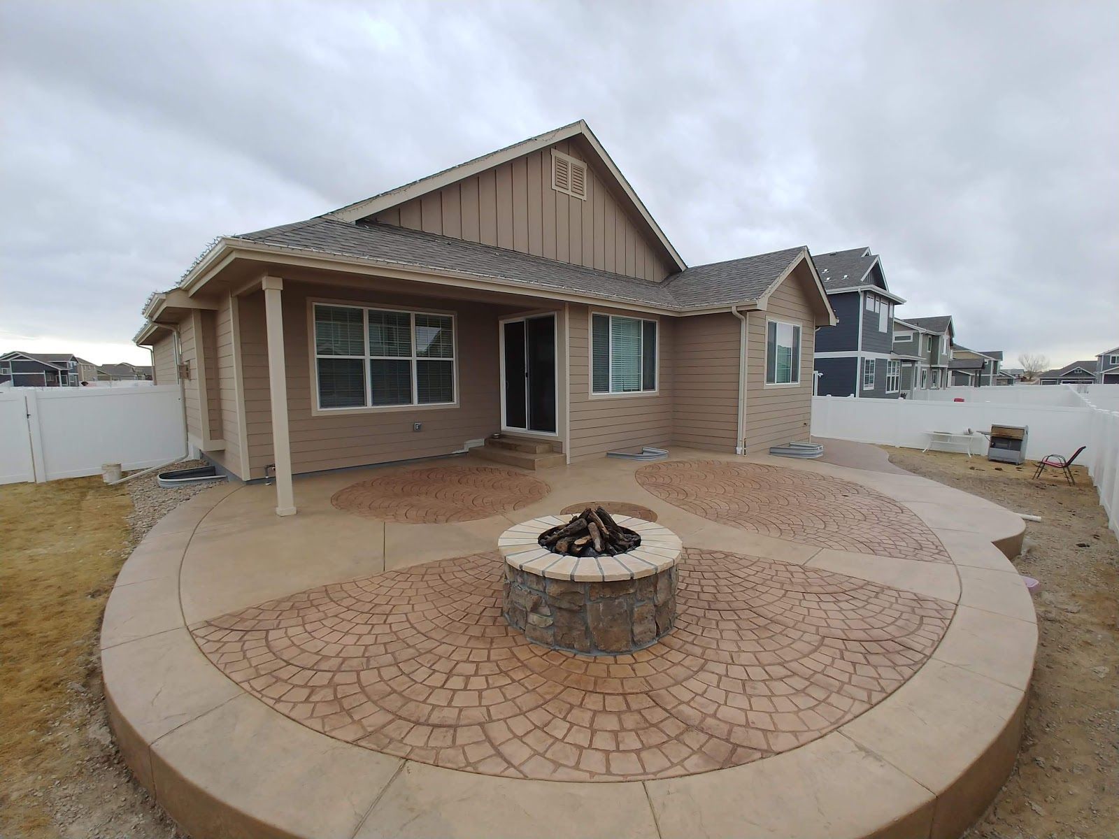 Backyard patio with fire pit, brown house, patterned concrete, white fence, cloudy sky.