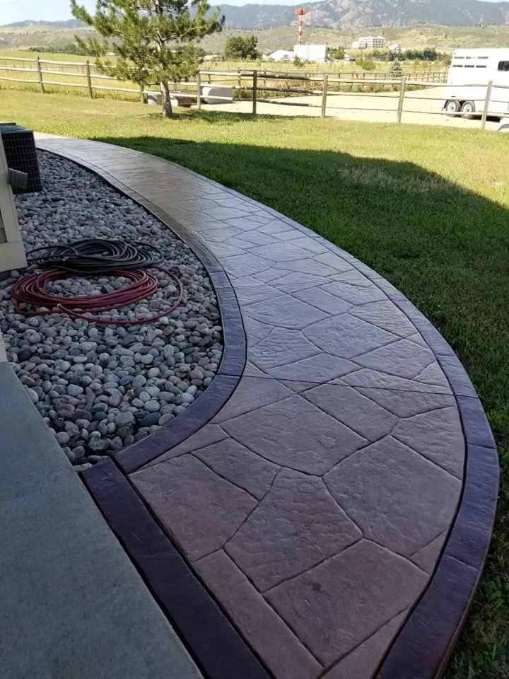 Curving stamped concrete walkway bordered by a dark edge, next to a rock bed and grass lawn.