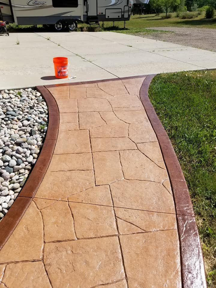 Stamped concrete pathway with brown borders and a stone-like pattern, next to a rock garden and grass.