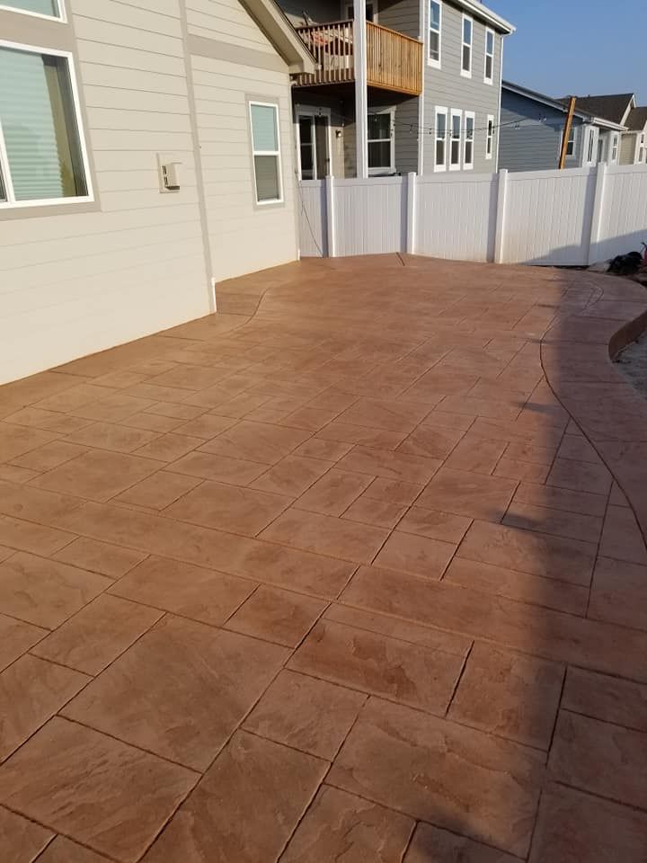 Stamped concrete patio with a brick pattern, brown color. Adjacent to a house and white fence.