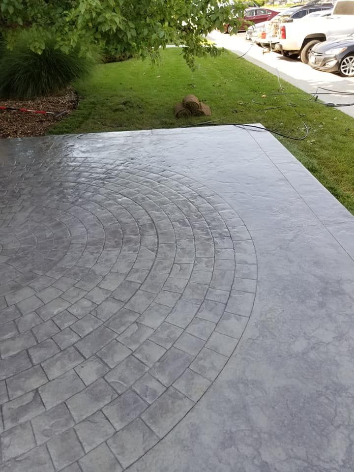 Gray stamped concrete patio with a curved brick pattern, next to green grass and a street with parked cars.