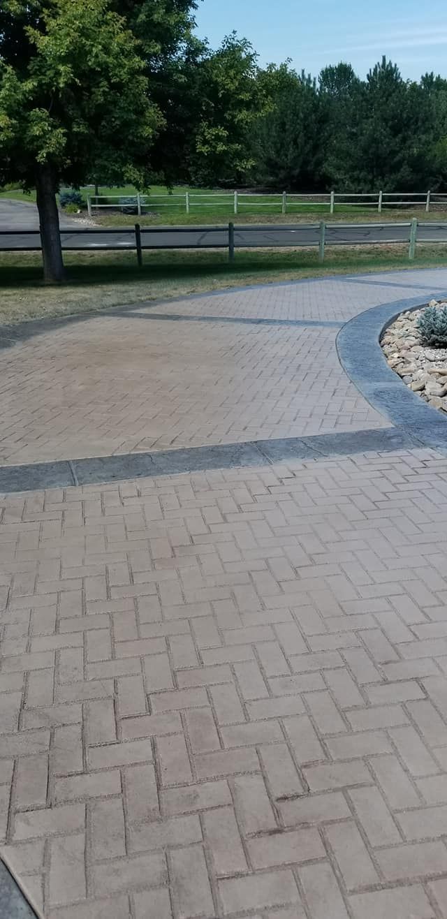 Brick patterned pathway leading to a grassy area, surrounded by trees and a fence.