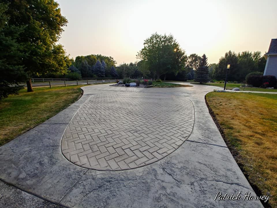 Concrete driveway with a brick pattern, curved design. Green grass, trees, and a sunset in the background.