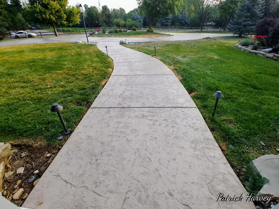 Concrete walkway curves through green lawn, flanked by garden lights, leading towards trees and houses.