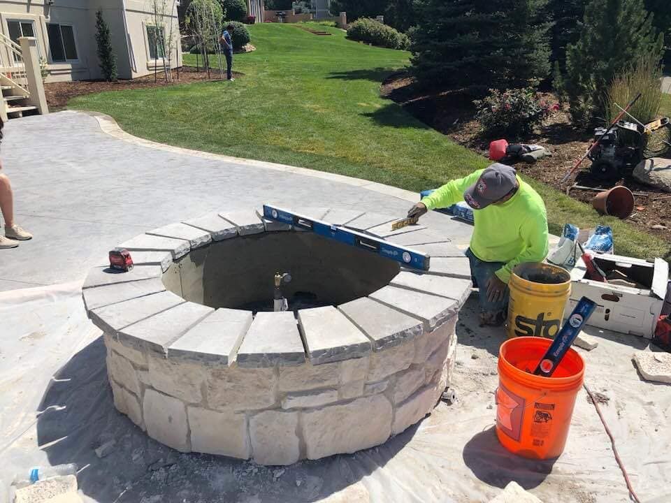 Man using a level to install stone around a fire pit. Sunny day, green grass, gray stone.