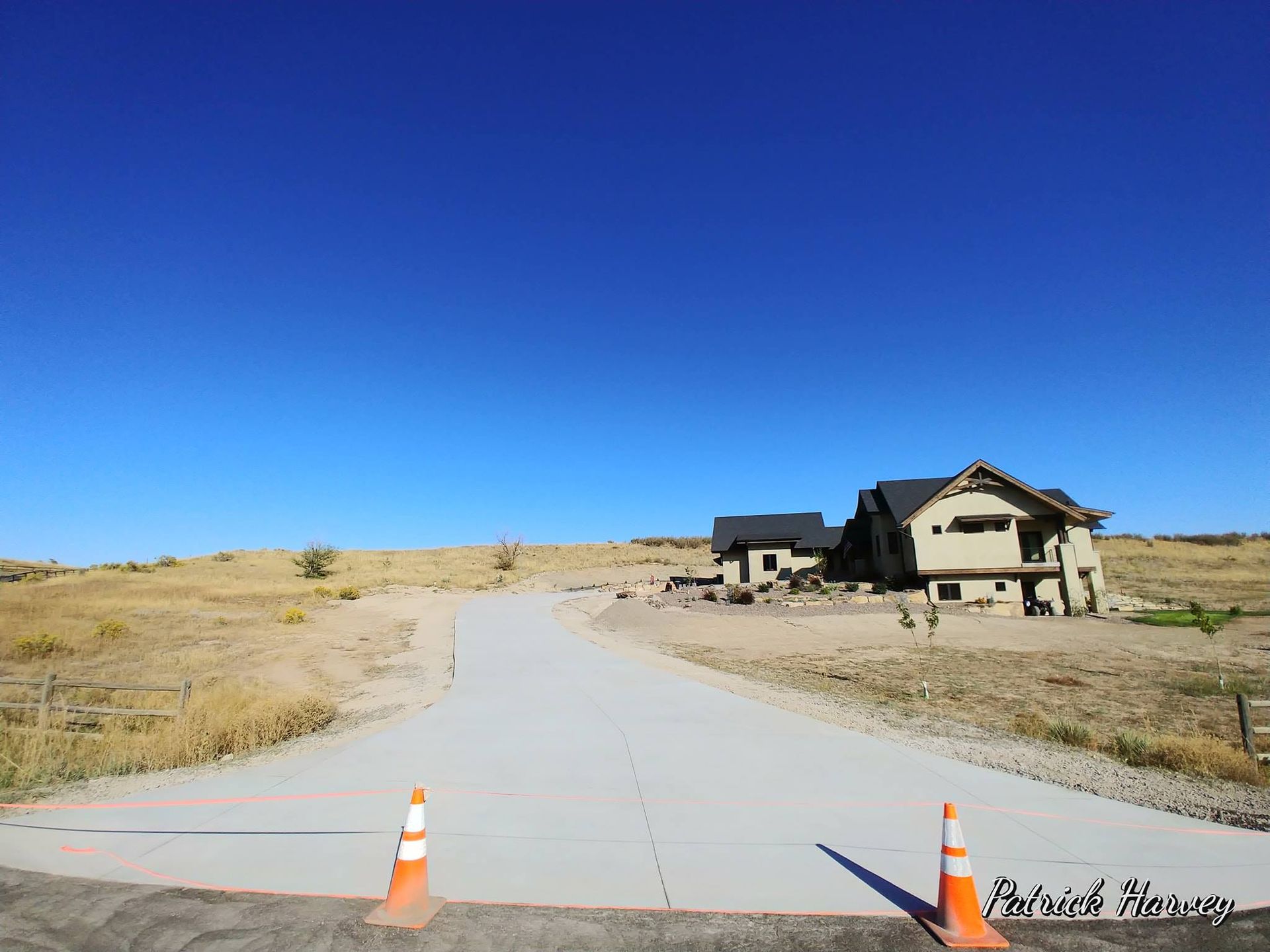 Paved road leads to unfinished houses under a clear blue sky. Dry landscape, orange cones in foreground.