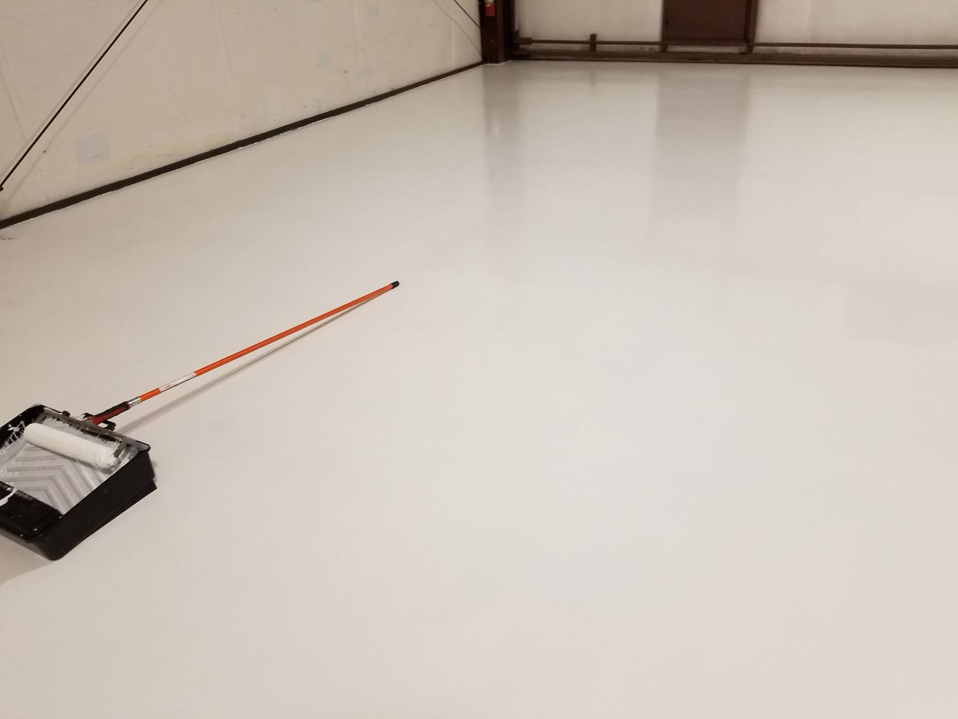 White epoxy floor being painted; roller and paint tray are on the floor in a large room.