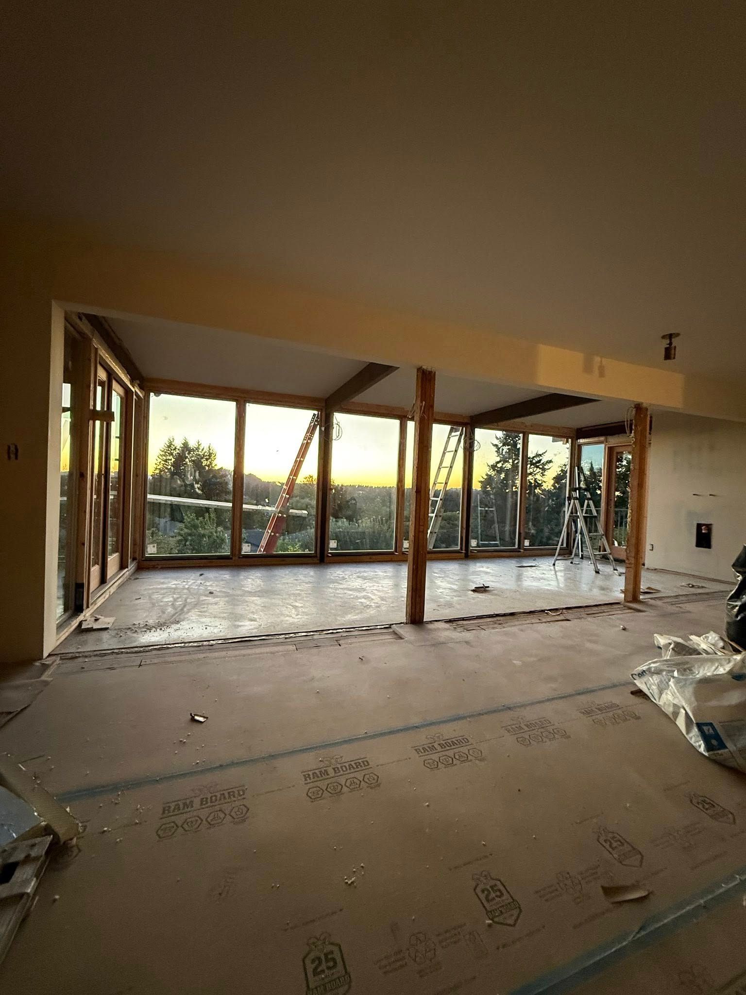 Interior of a room under construction with large windows overlooking a sunset. Wooden supports are visible.