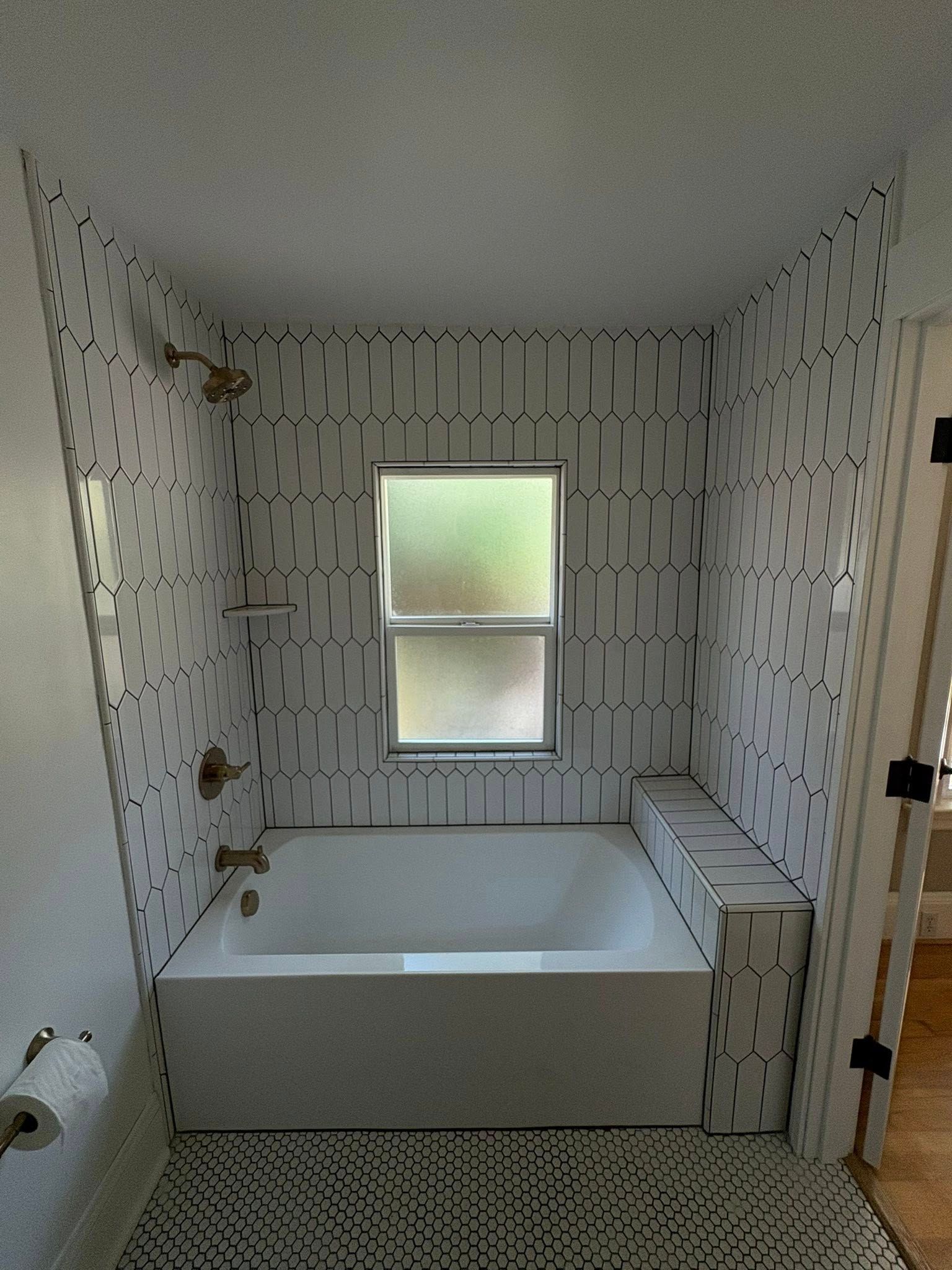 Bathroom with white bathtub, geometric tile, window, and gold fixtures.