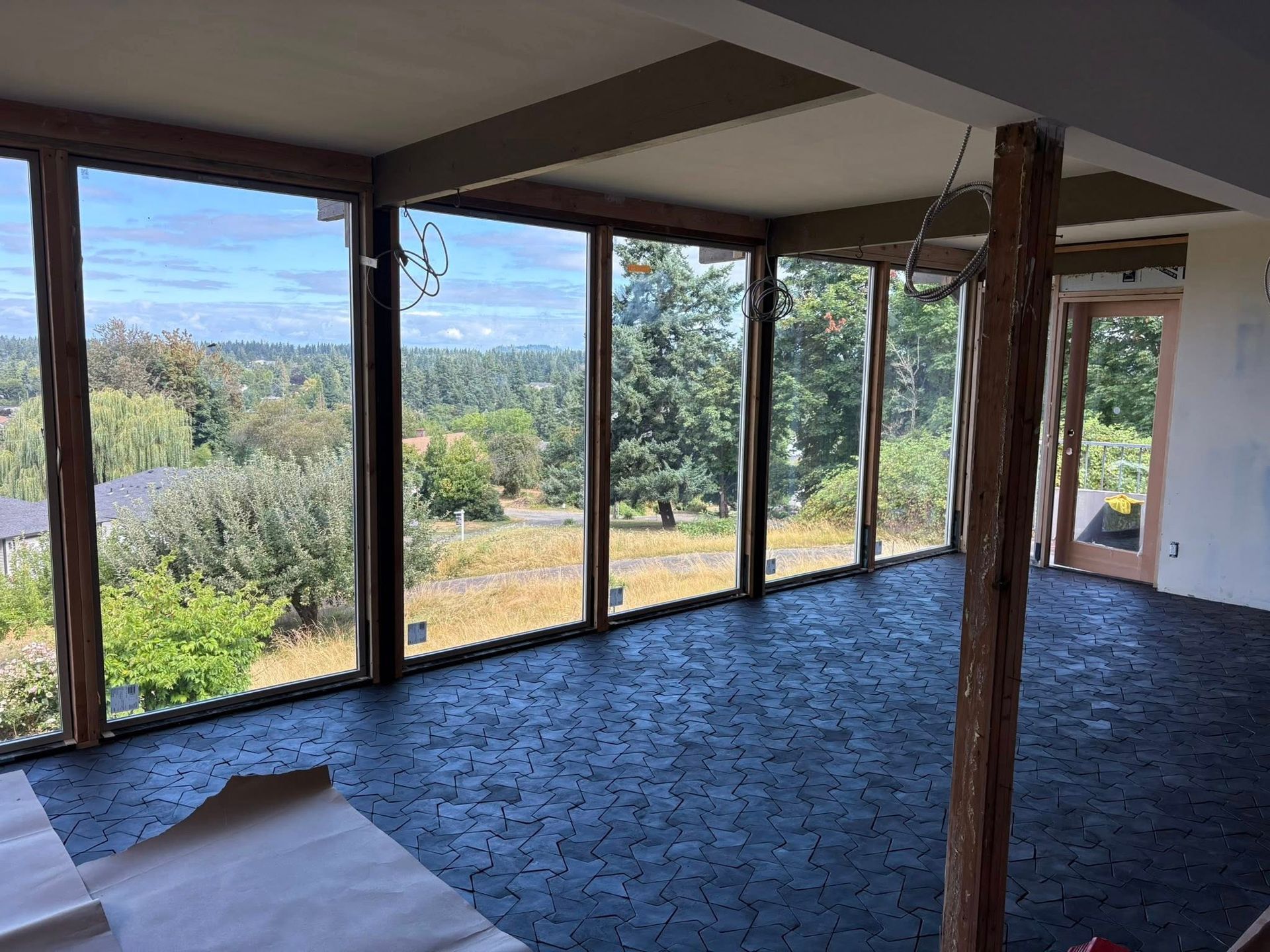 Sunroom with large windows, hexagonal dark floor tiles, wooden support beams, and a view of green trees and sky.