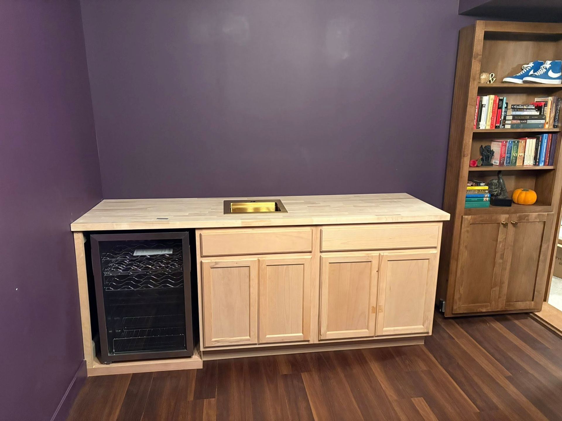 Built-in bar with a mini fridge, cabinets, a sink, and a wooden countertop against a purple wall, with a bookshelf to the right.