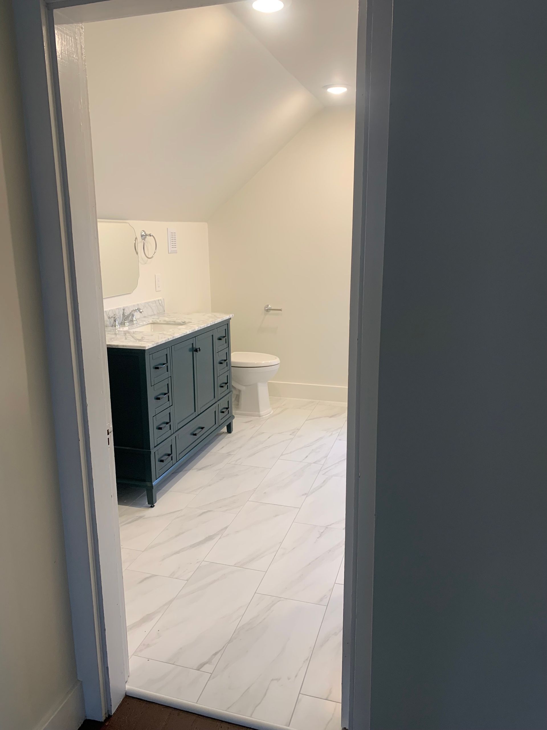 Bathroom interior: blue vanity, white toilet, marble floor, and recessed lighting visible through doorway.