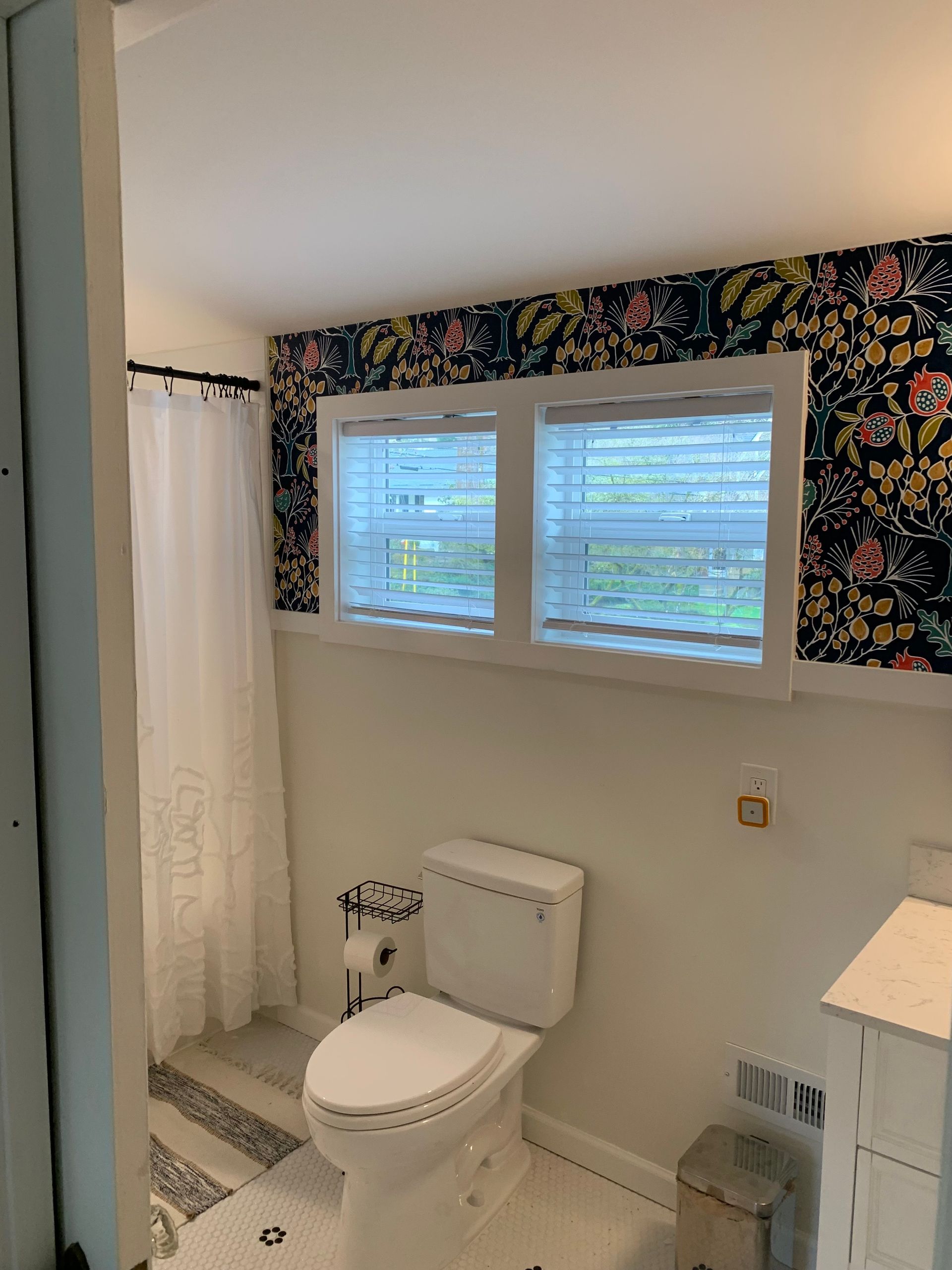 Bathroom with floral wallpaper, toilet, windows with blinds, and a white shower curtain.