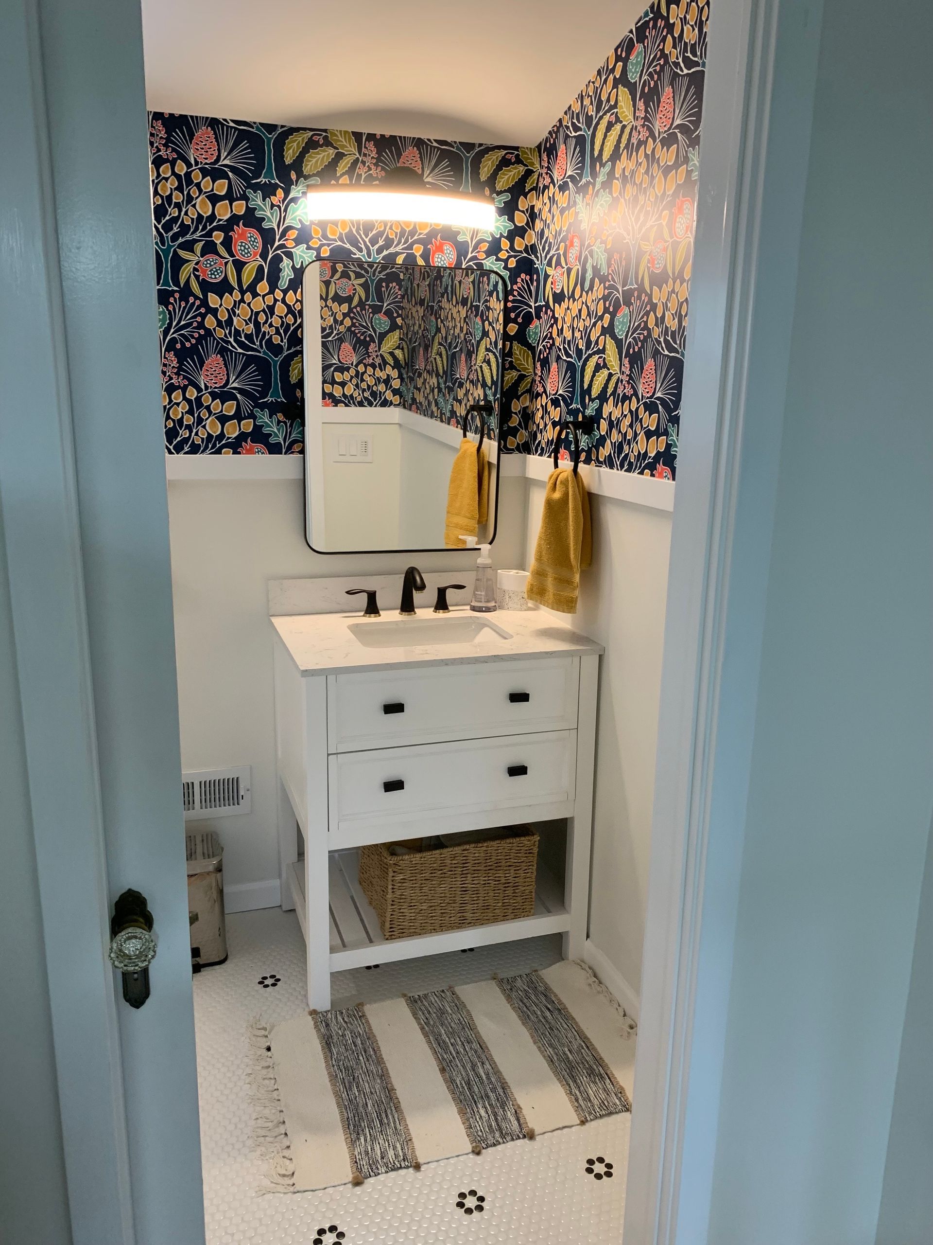 Small bathroom with floral wallpaper, white vanity, black faucet, and striped rug.