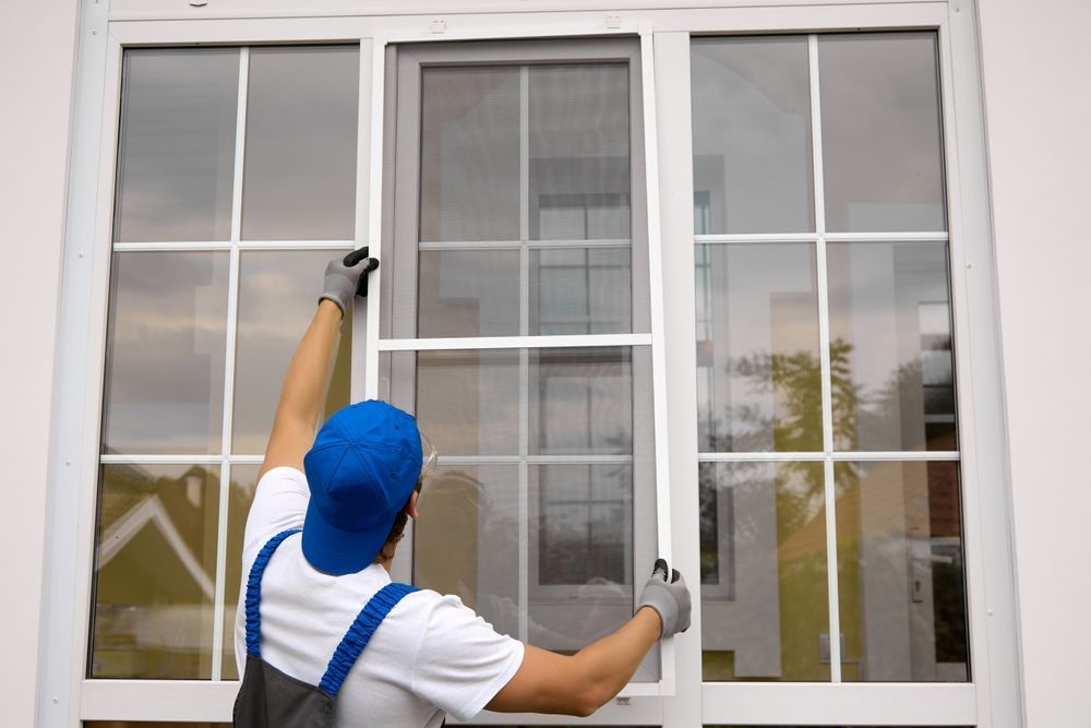 Person in blue cap and work clothes installing a white window screen on a white building.