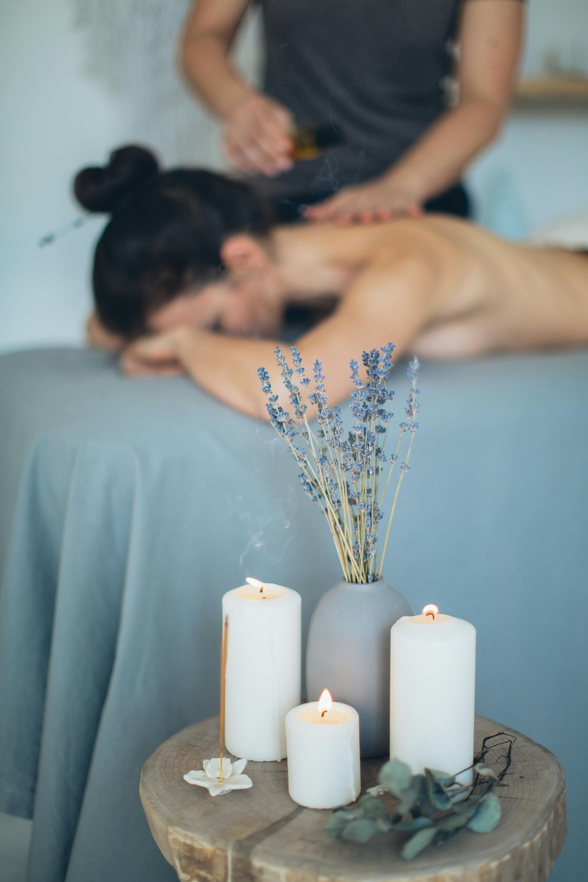 en arrière plan un massage fait un massage bien-être à une femme.
En premier plan des éléments de décoration avec des bougies et des fleurs sur un tabouret en bois. 
Massage bien être, massage relaxant, massage détente, colmar, ribeauvillé, horbourg wihr, wintzenheim