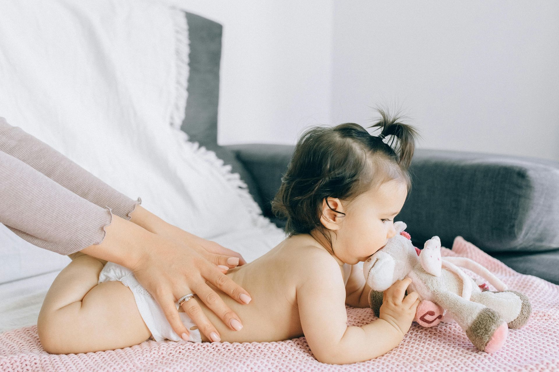 une petite fille est allongée sur le ventre sur un lit, avec une peluche entre les mains. Des mains de femme la tienne à la taille. 
Massage bébé, yoga bébé, Colmar, Ribeauvillé, Wintzenheim, Horbourg Wihr