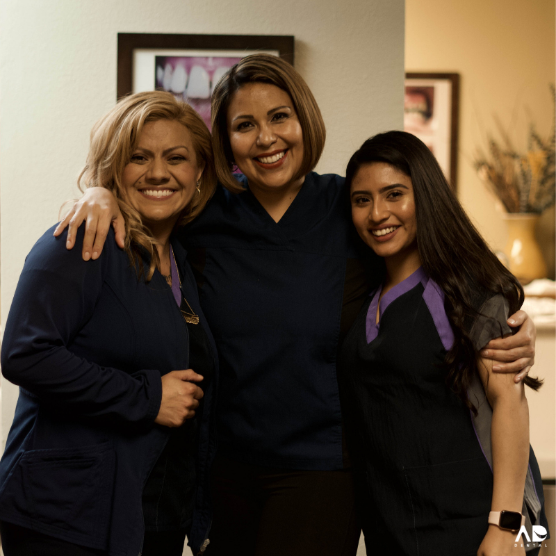 Three women in medical scrubs smile, with arms around each other in a warmly lit office setting. Three women in medical scrubs smile, with arms around each other in a warmly lit office setting.