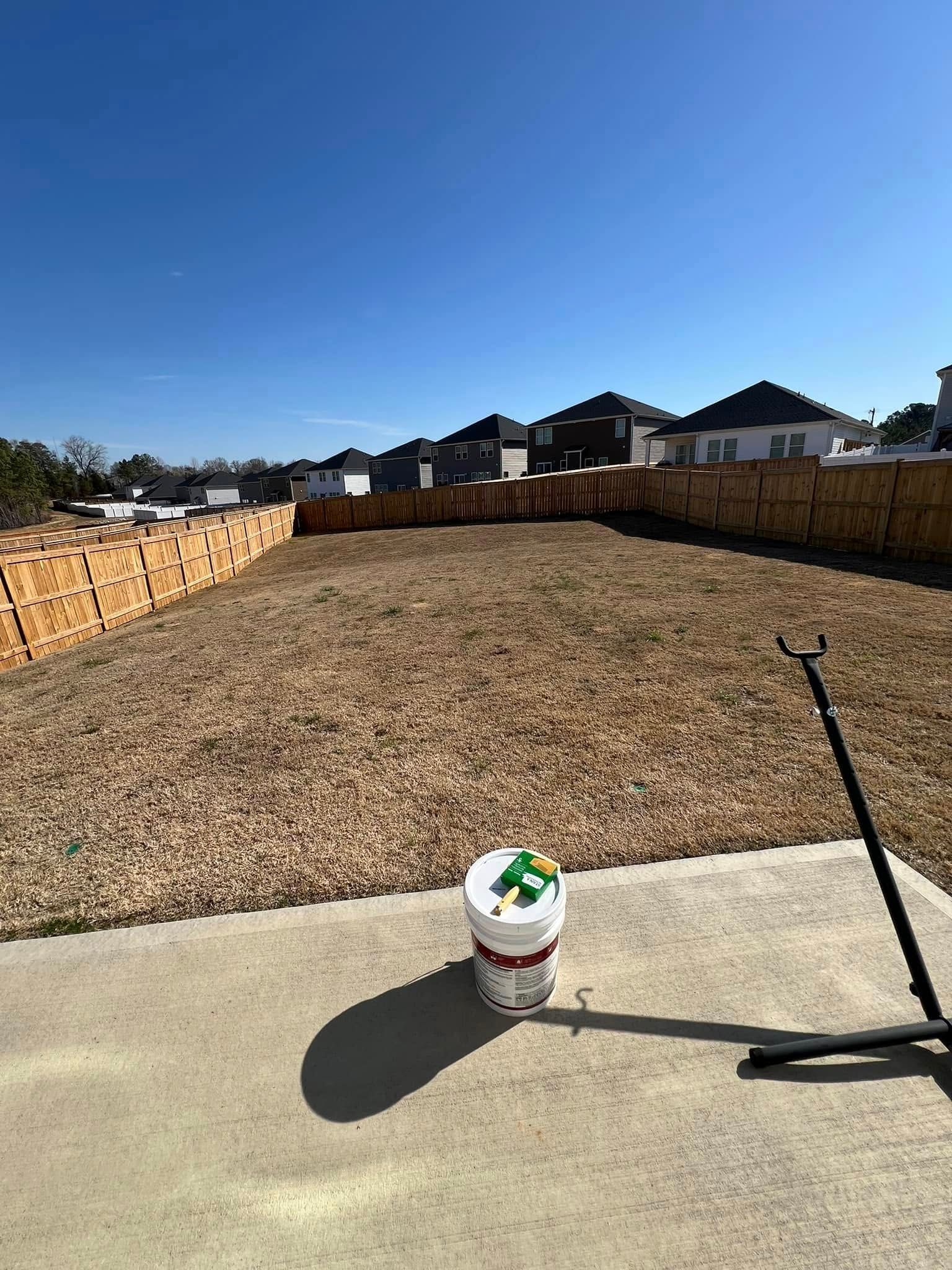 A dry lawn enclosed by a wooden fence, a bucket and a metal stand on a concrete patio under a clear blue sky.