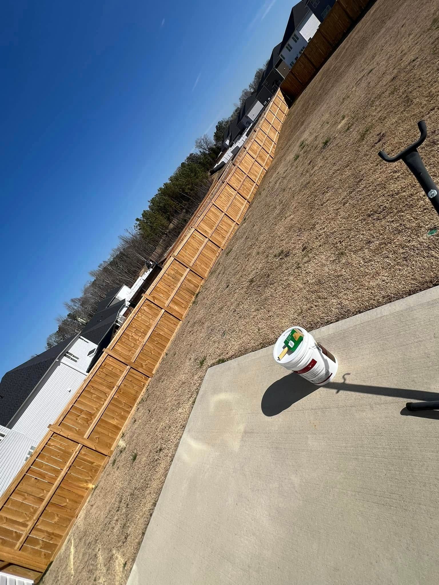 Wooden fence along a hillside with gravel, blue sky overhead, bucket and hose on a concrete pad.