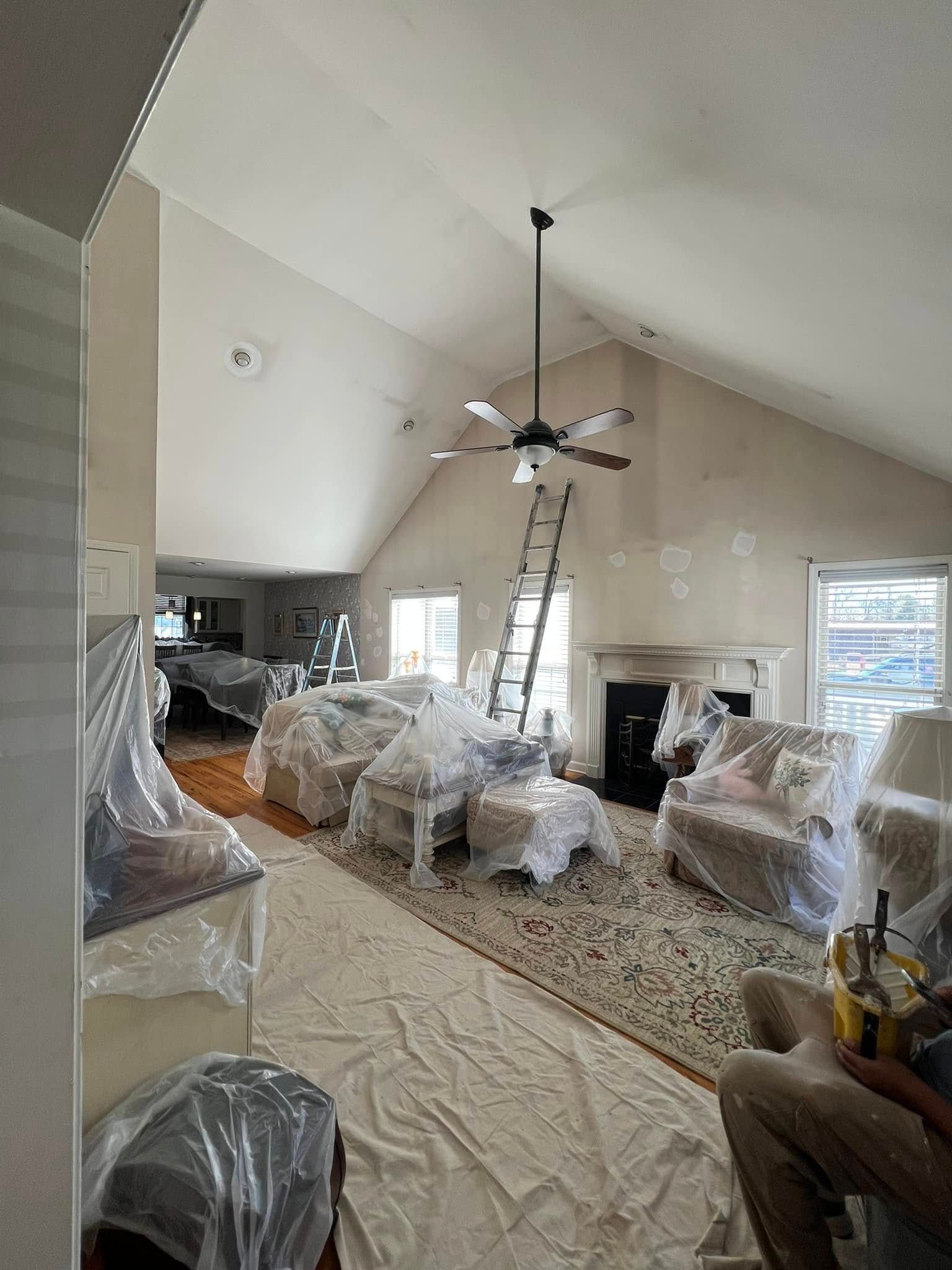 Living room being painted, furniture covered in plastic, ladder, high ceiling, fireplace.