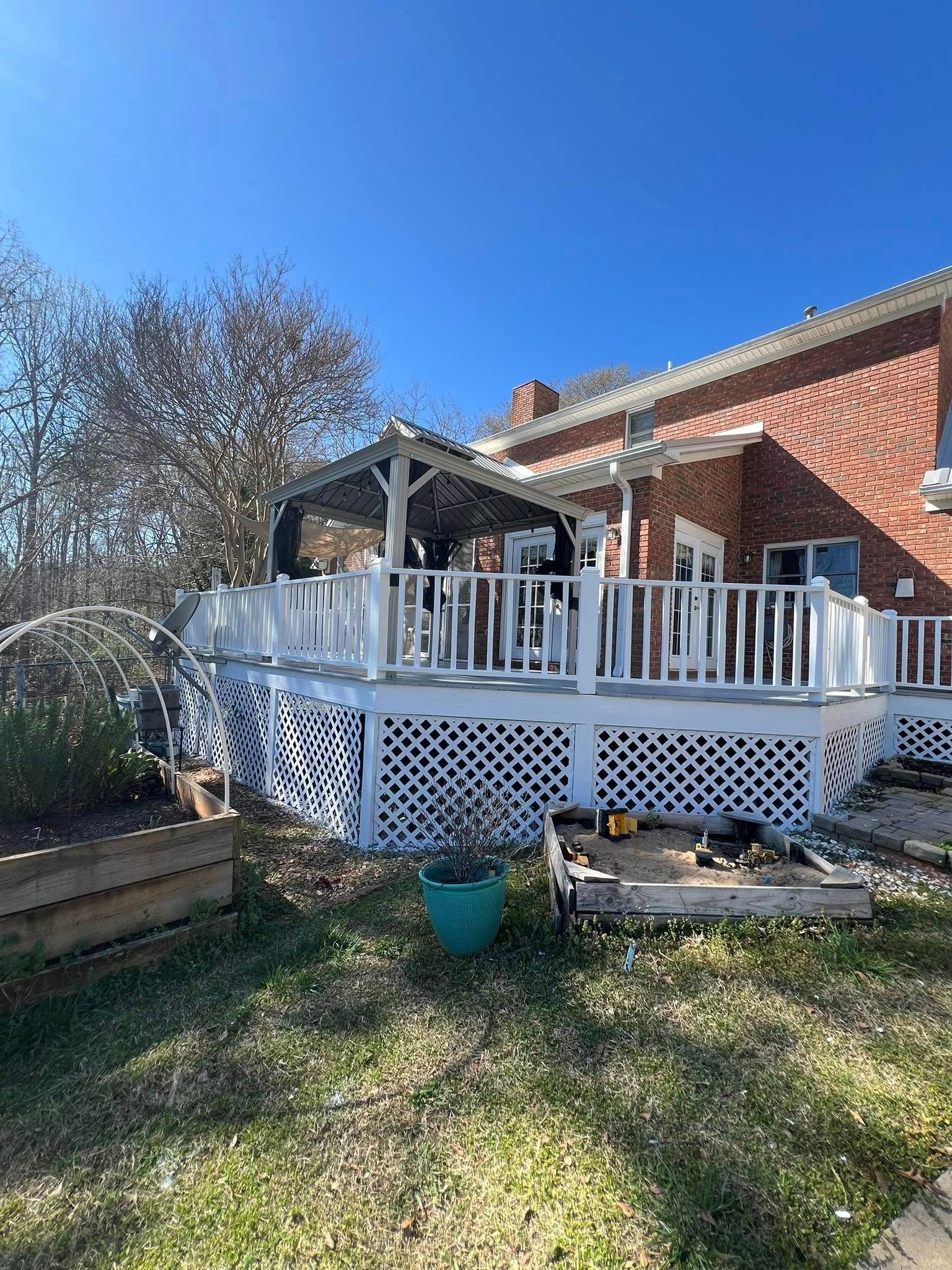 White deck with lattice skirt, pergola, and brick house under a blue sky.