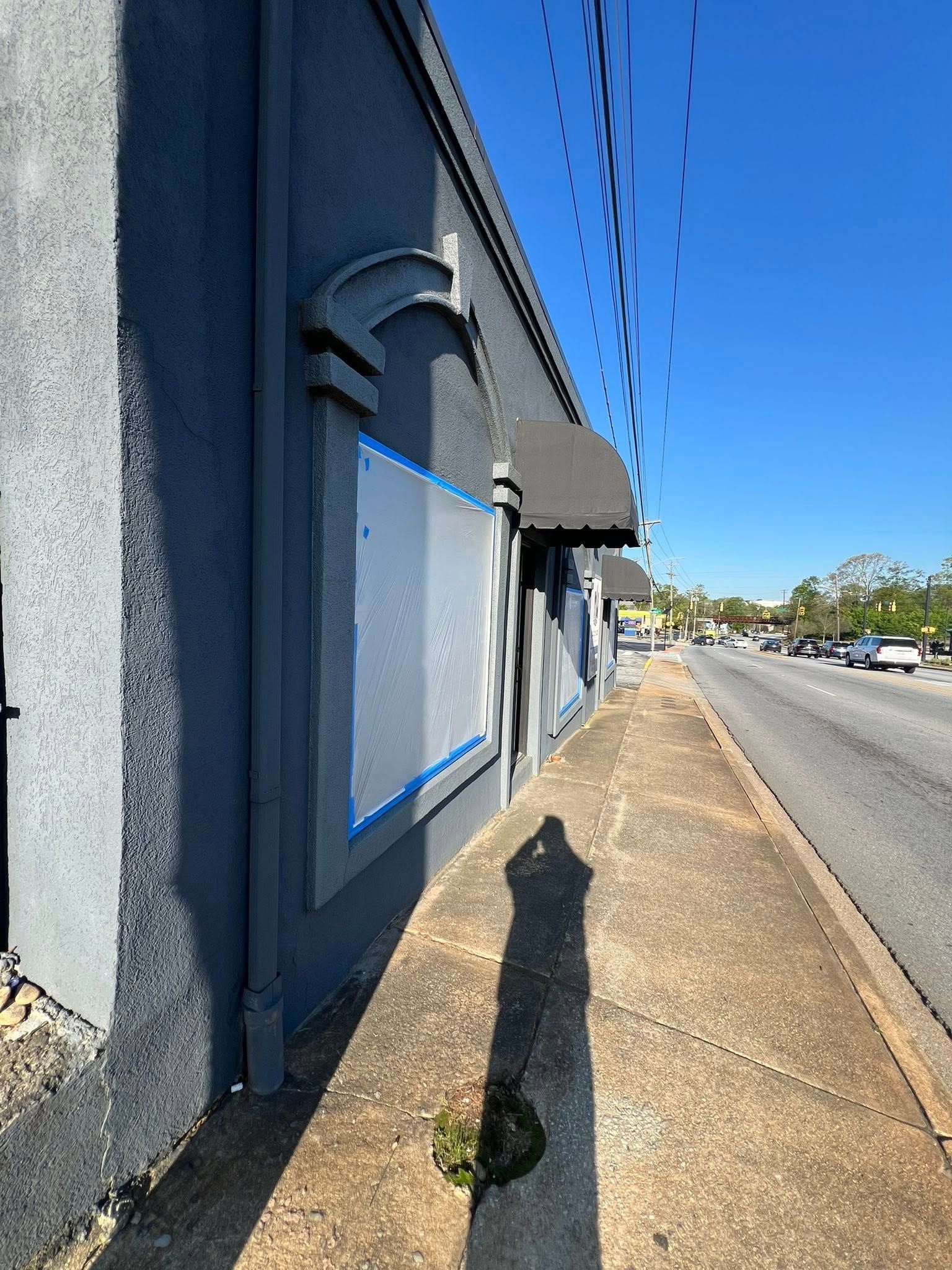 Gray building with a sidewalk, road, and a shadow cast by the sun on a sunny day.