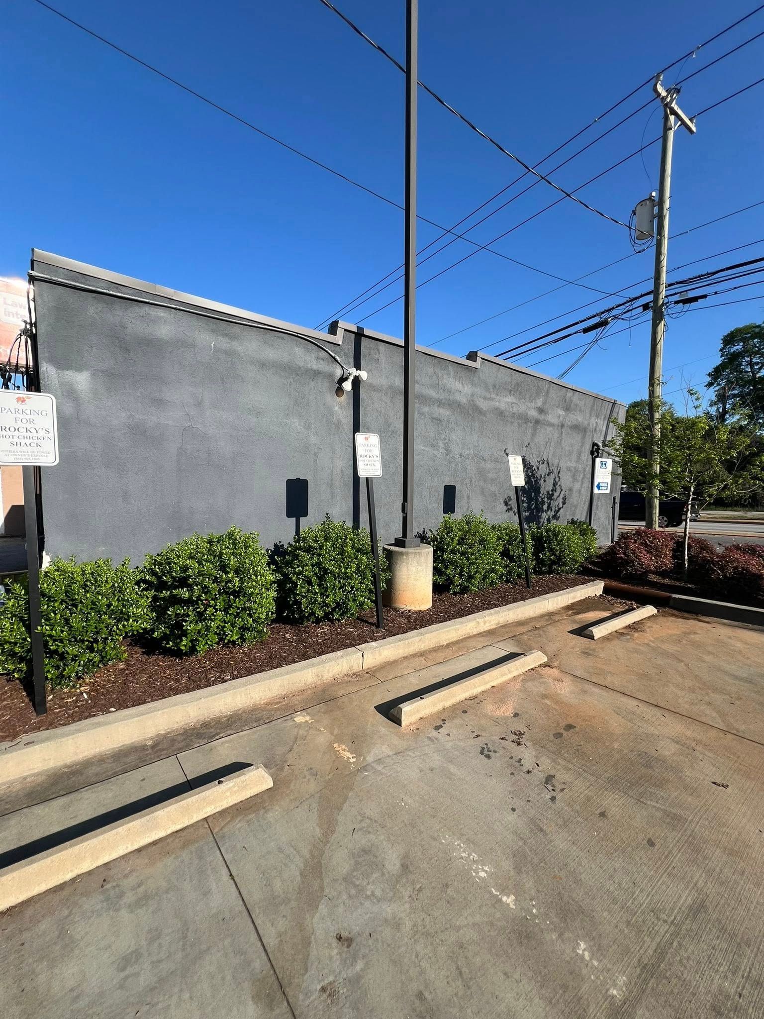 Exterior of a gray building with bushes and parking space. Clear blue sky and overhead power lines.