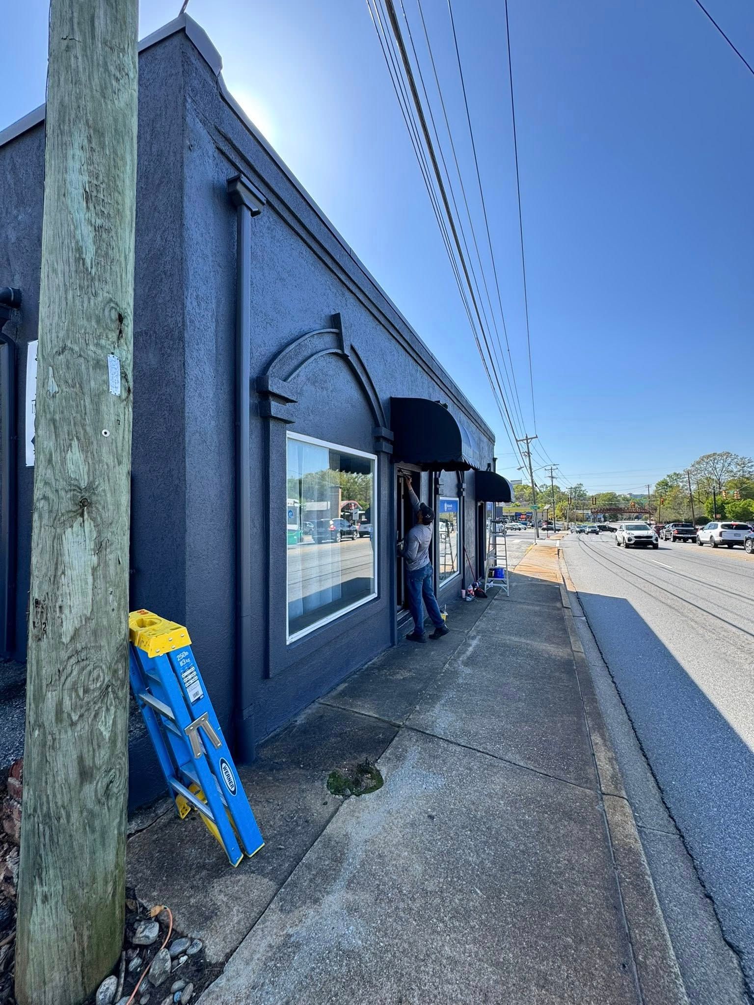Dark blue building with awning. Person stands in doorway. Ladder leans on utility pole on sidewalk.