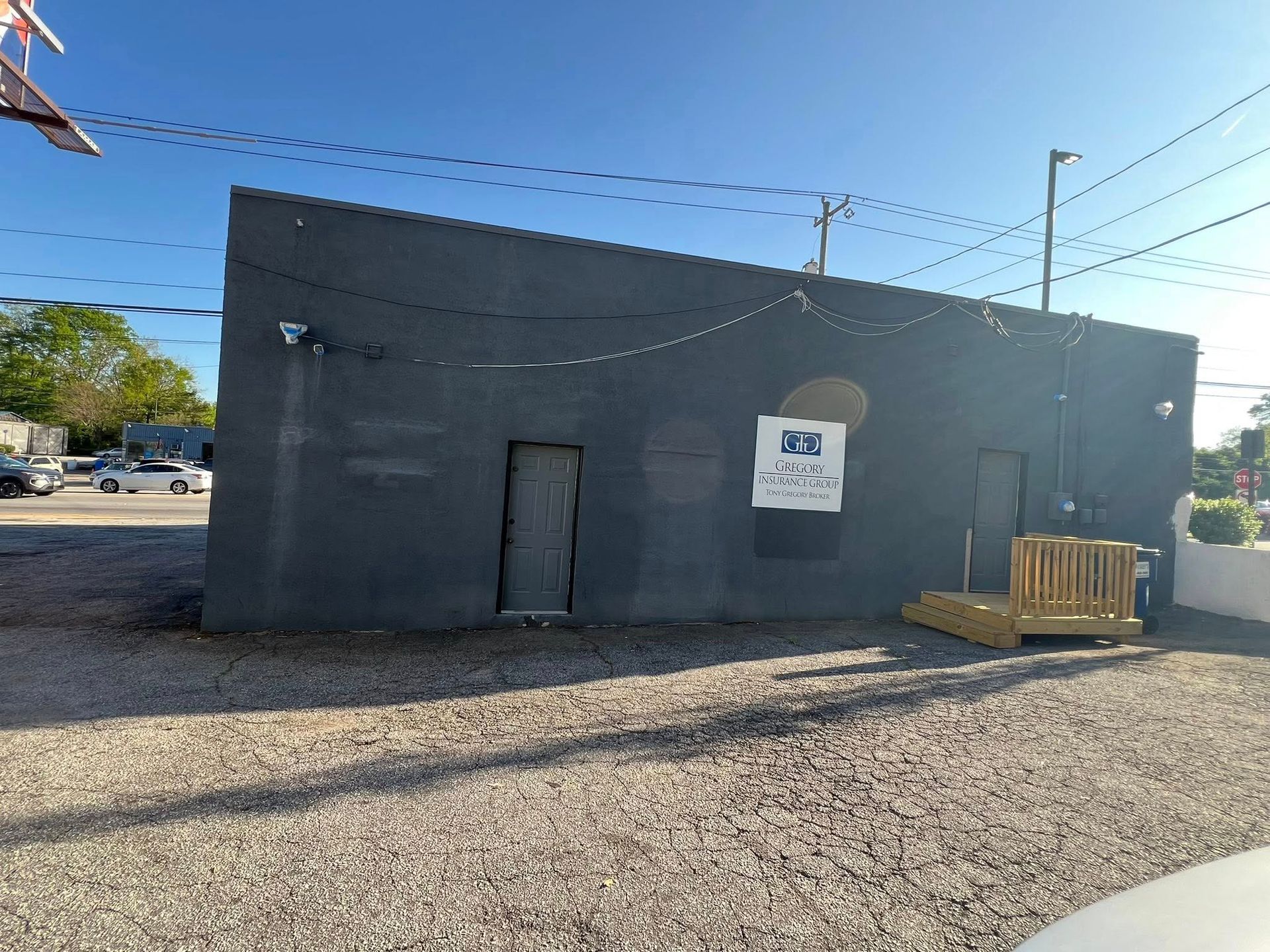 Gray building with two doors, a sign, and a wooden platform, set on a gravel lot under a blue sky.