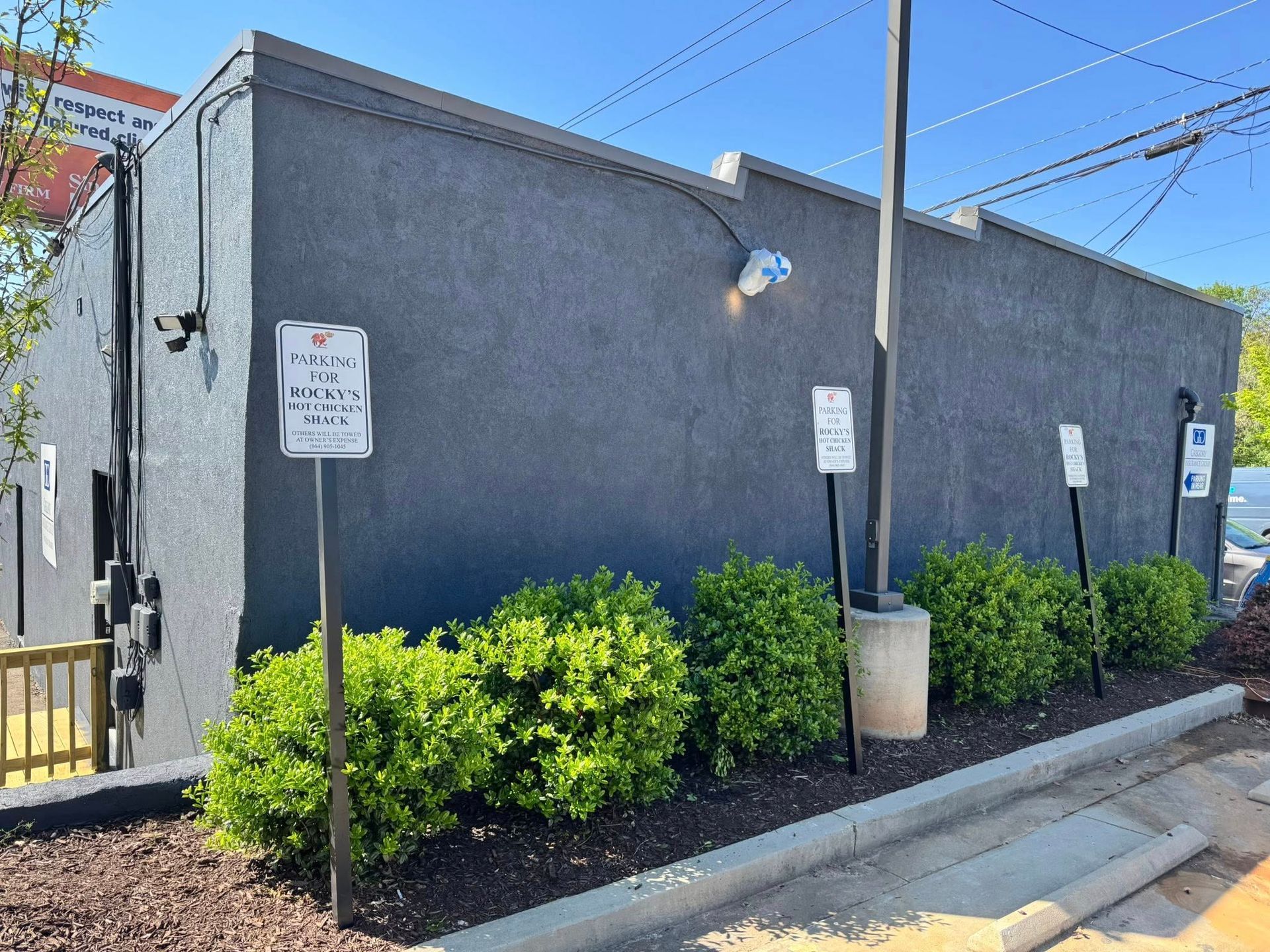 Exterior of a dark gray building with parking signs, bushes, and a light-colored curb.
