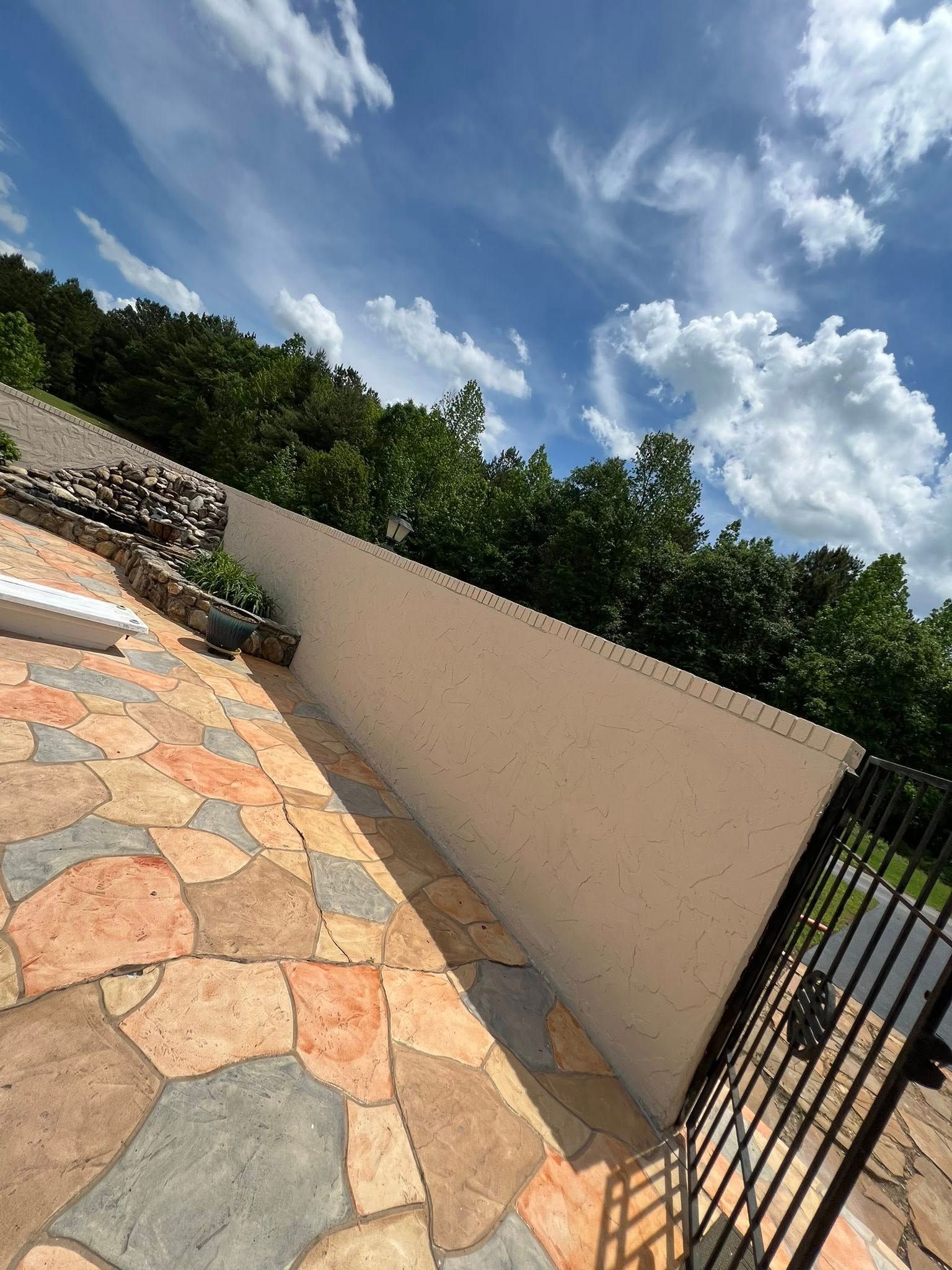 Patio with stone floor and white wall, trees, and a blue sky with clouds.