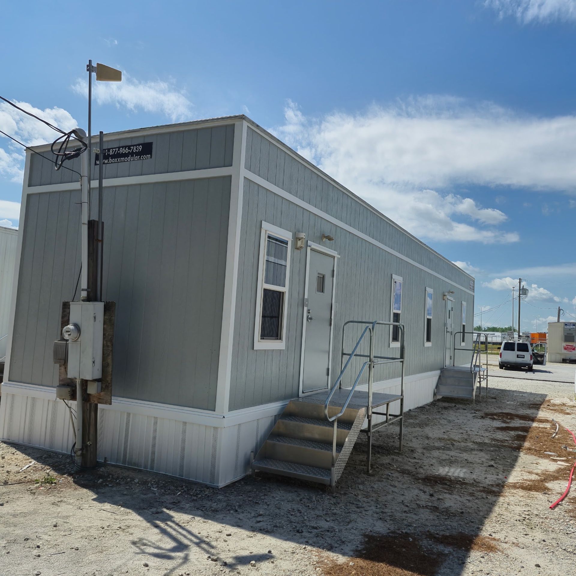 Gray modular building with stairs, a door, and windows on a construction site under a blue sky.