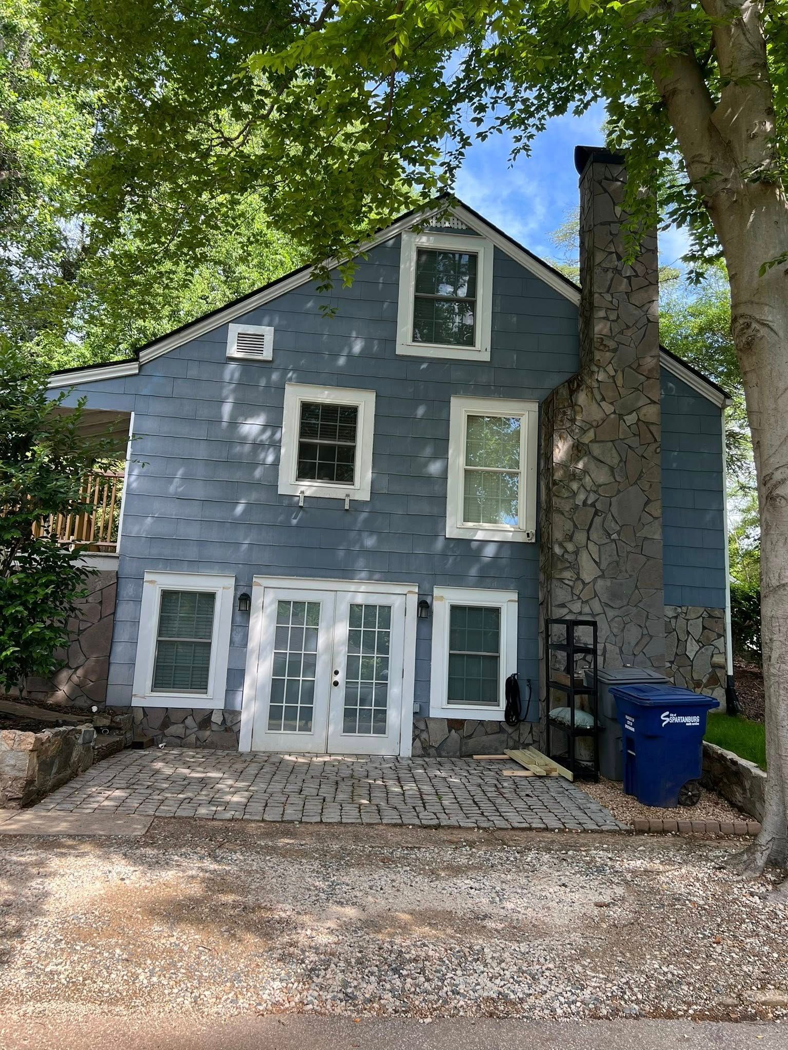 Blue-sided house with white-framed windows and doors, tall stone chimney. A blue trash can sits beside it.