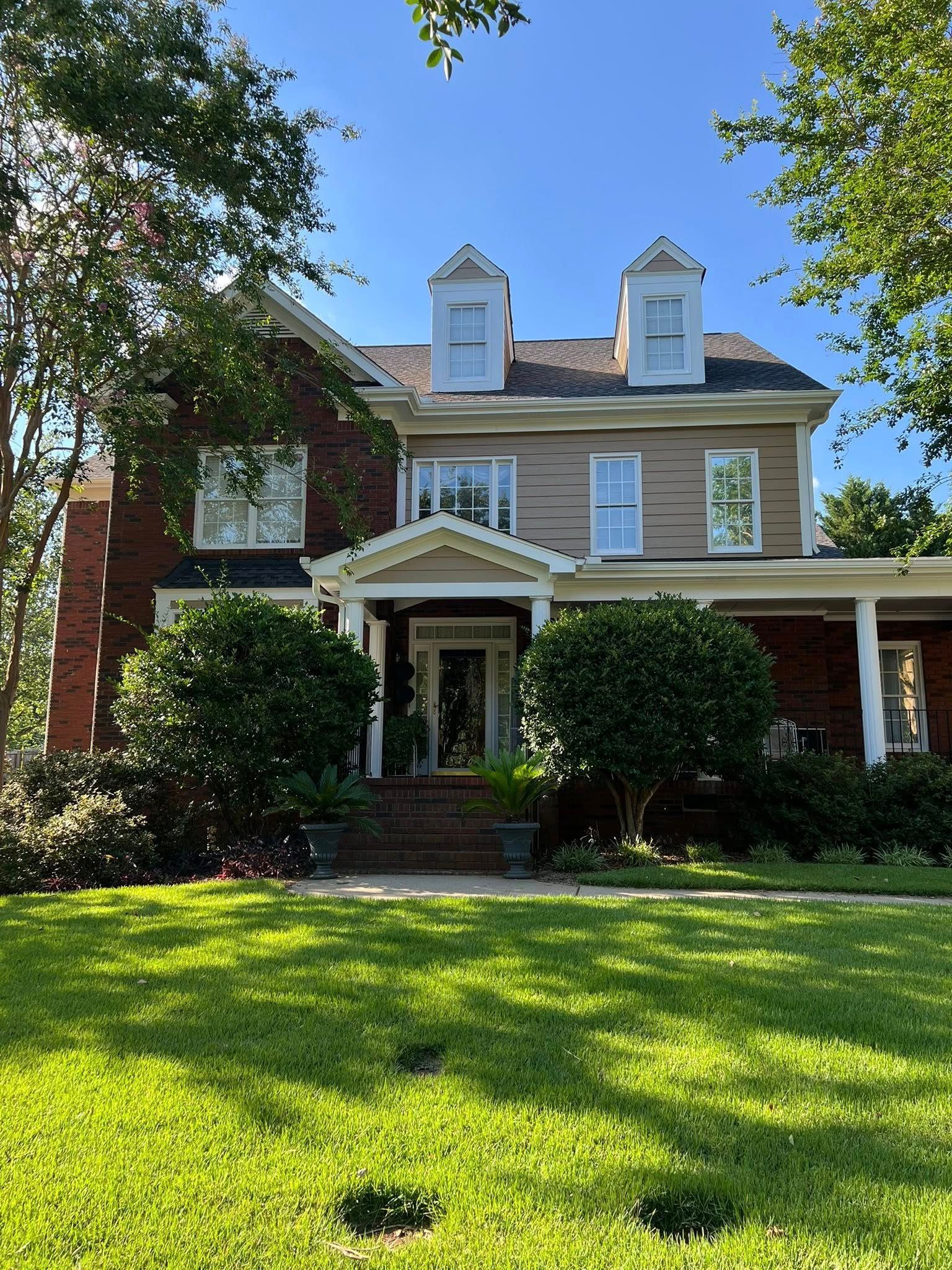 Large two-story house with brick and tan siding, covered porch, green lawn, and blue sky.