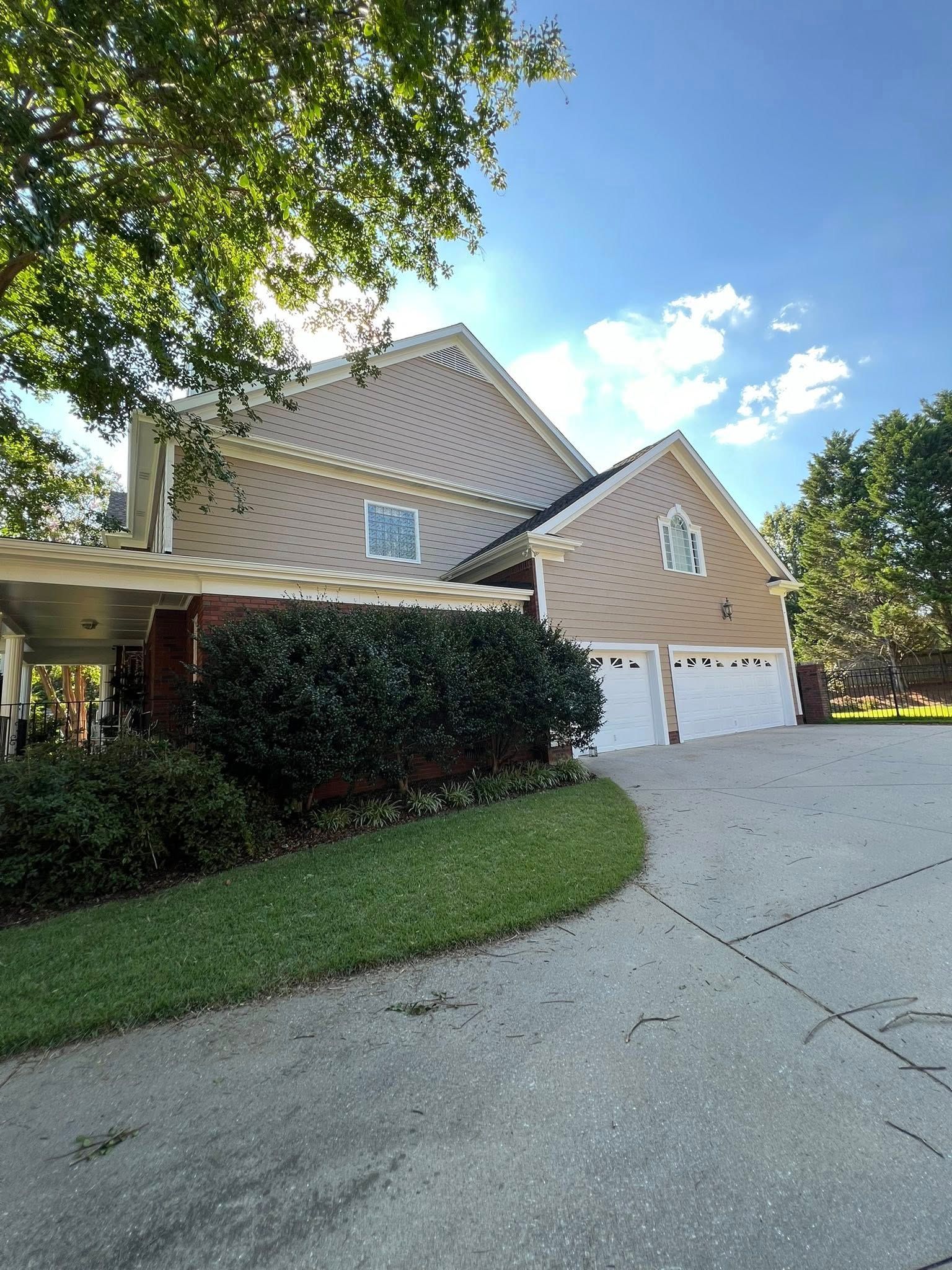 Two-story tan house with white garage doors, driveway, and surrounding greenery under a partly sunny sky.